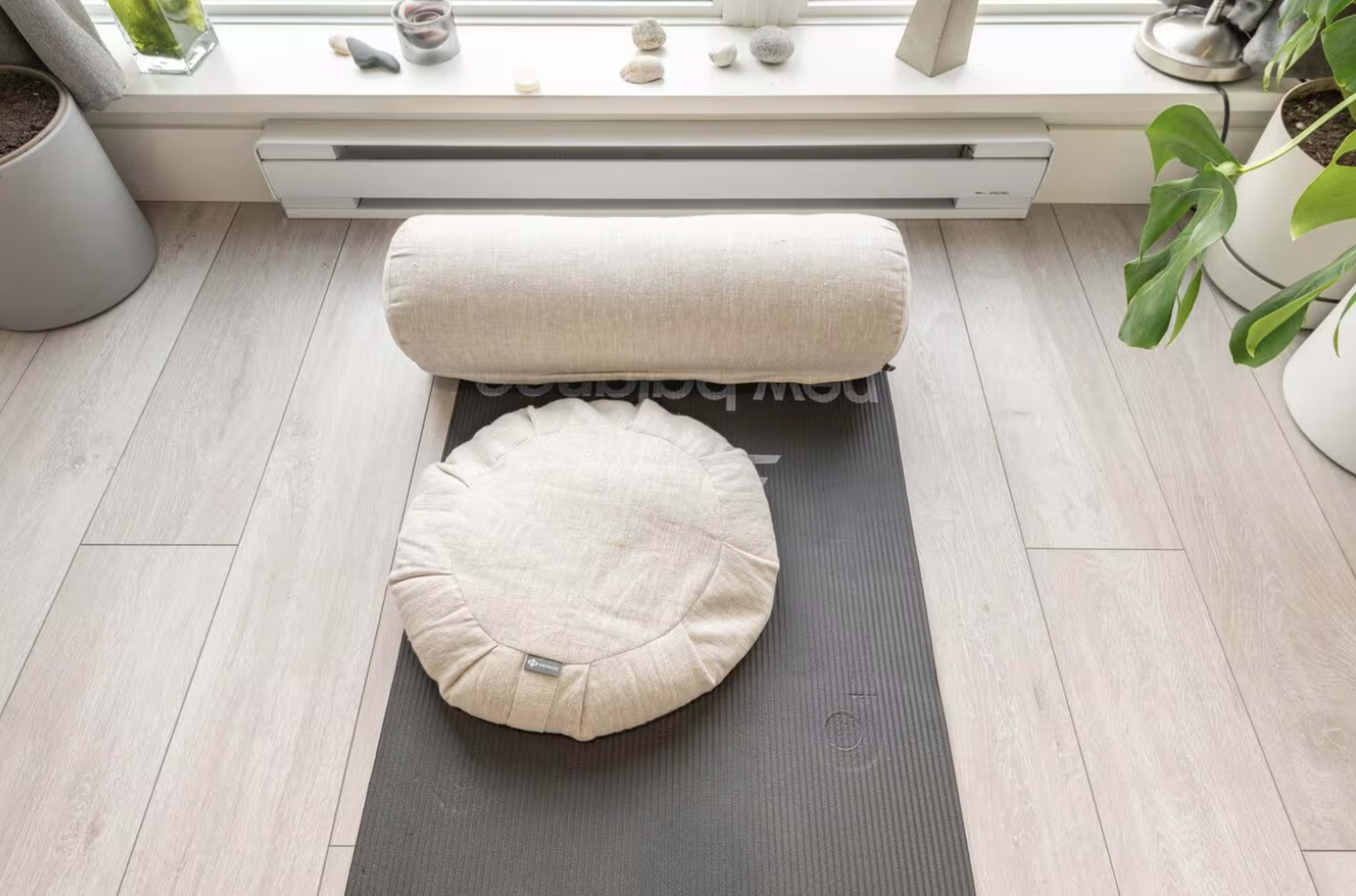 A yoga mat with a cylindrical and a round cushion placed on it is positioned near a window.
