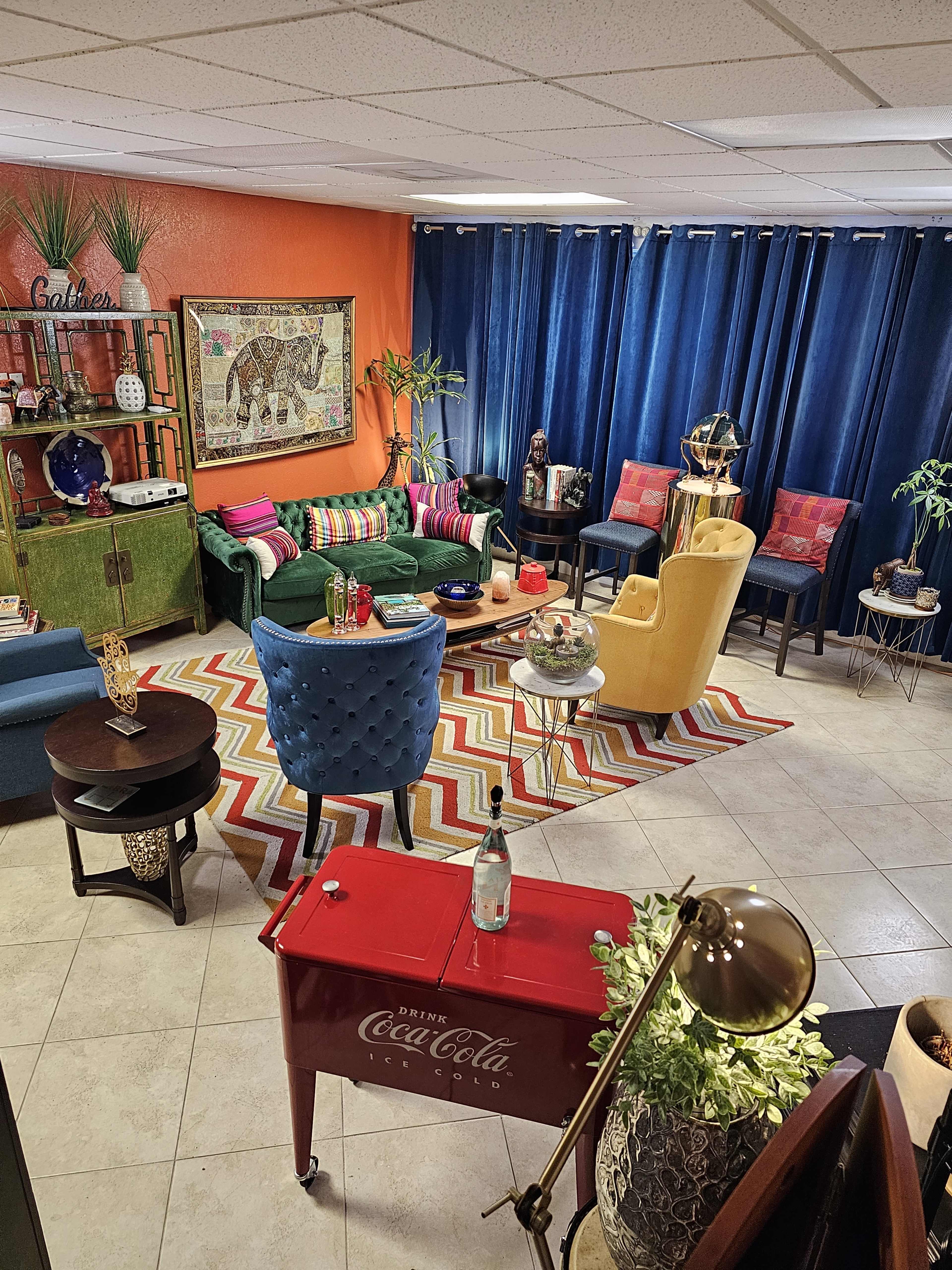 A brightly colored living room features a mix of furniture, including a green sofa, blue chairs, and a red Coca-Cola table, arranged around a patterned rug.
