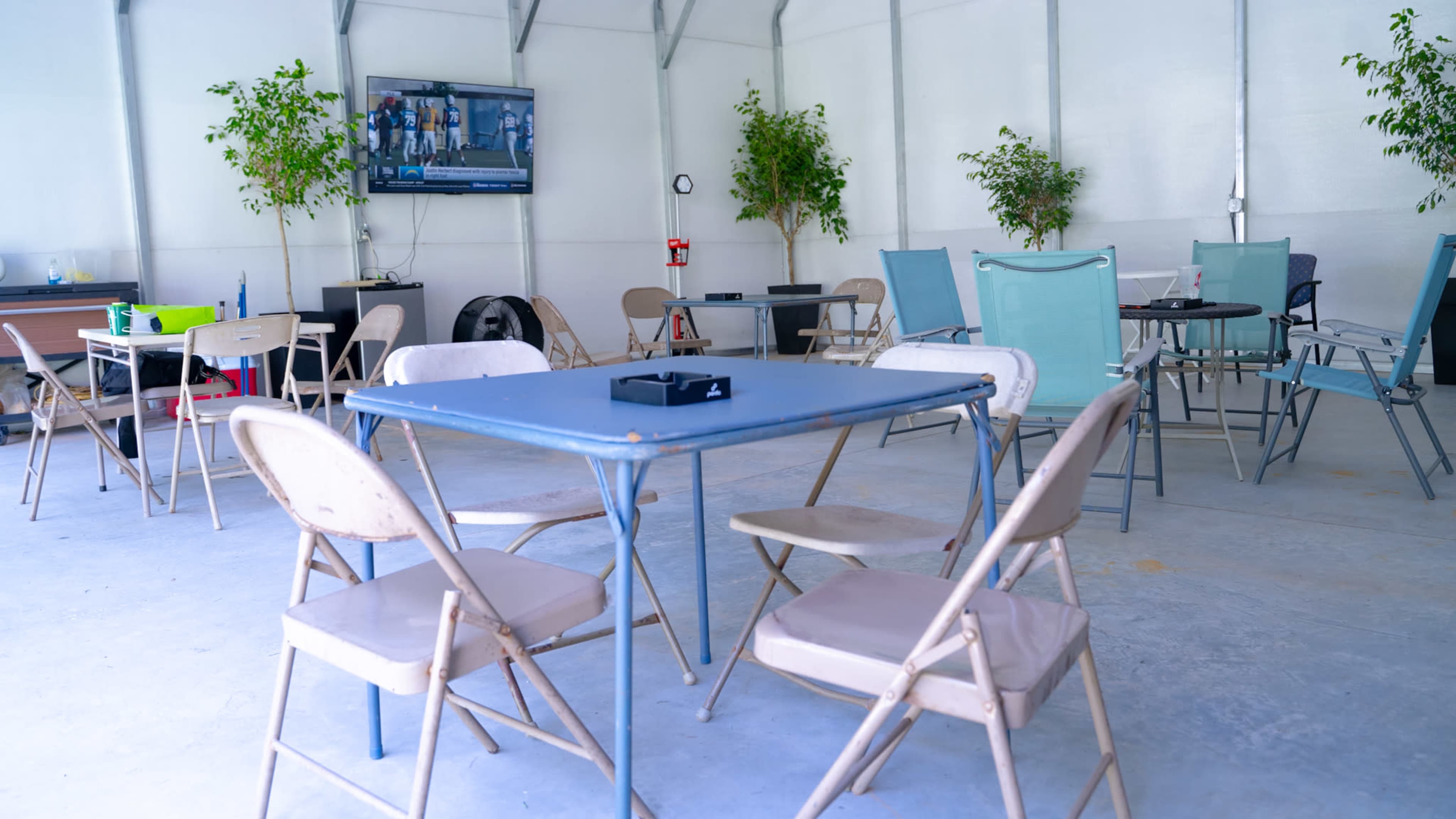 A spacious indoor area with multiple tables and chairs arranged around, featuring a television mounted on the wall and potted plants in the corners.