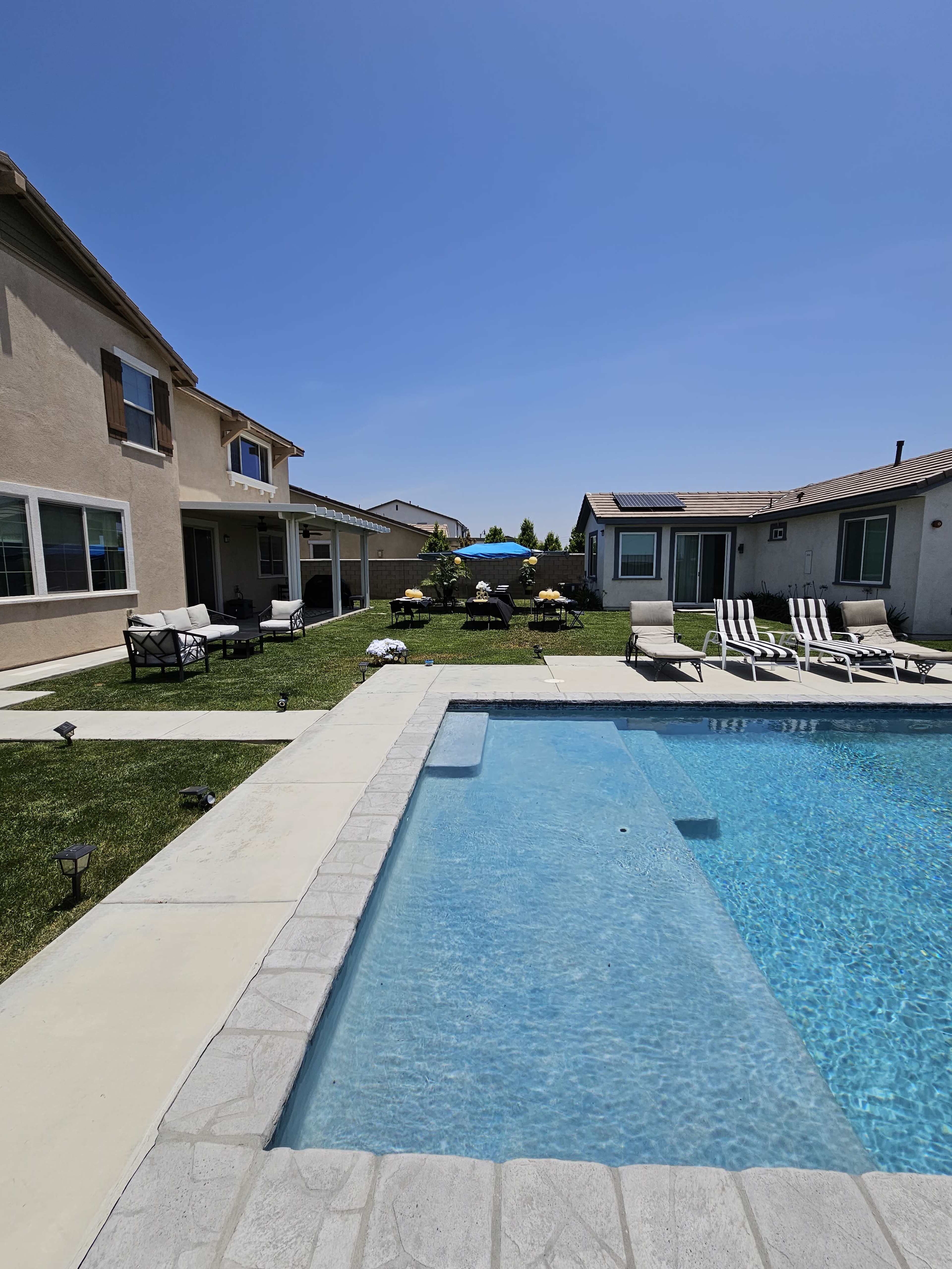A backyard pool area features a clear swimming pool surrounded by lounge chairs, with two houses and well-maintained grass in the background.