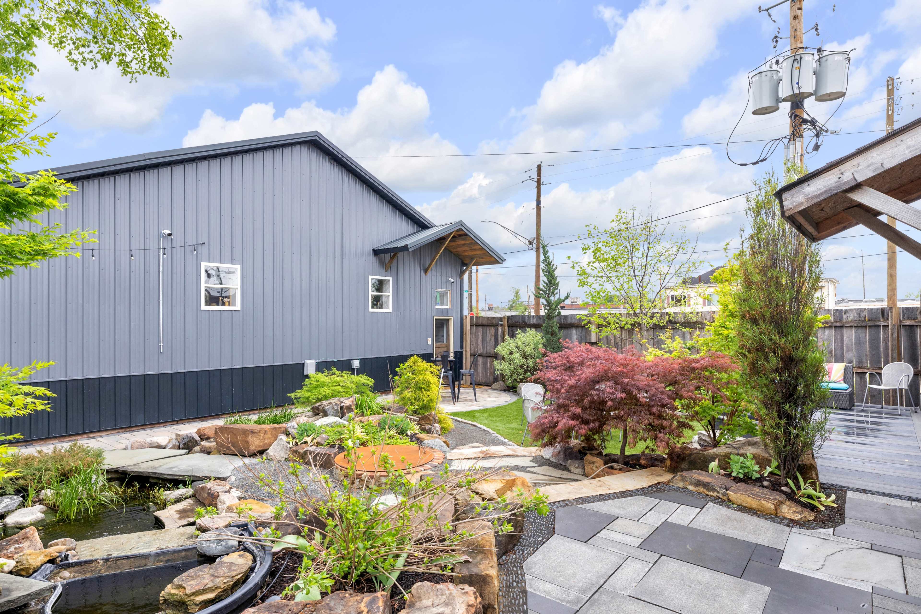 The image shows a landscaped backyard featuring a pond surrounded by rocks, plants, and a pathway leading to a gray building.