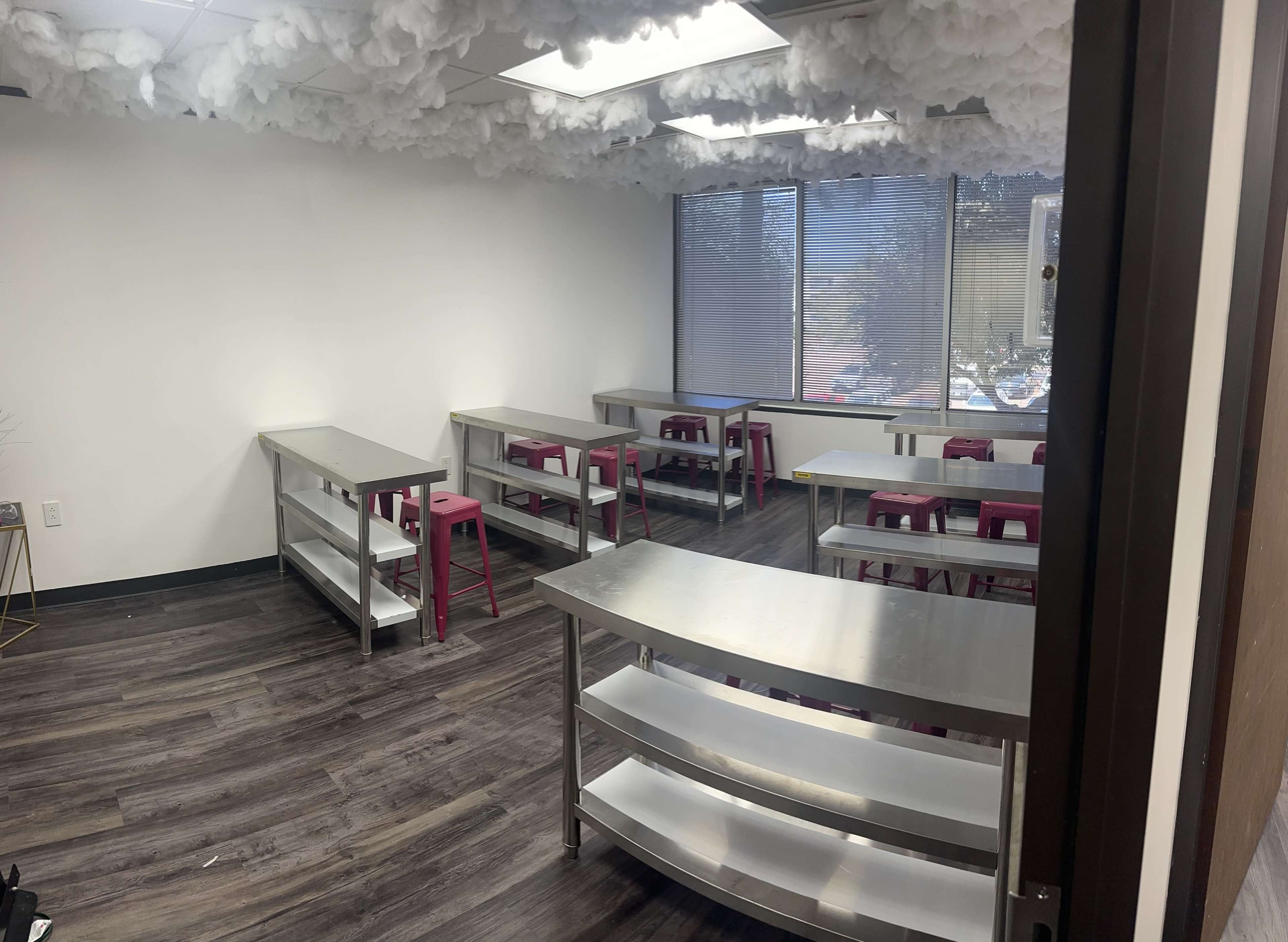 A classroom with several metal tables placed in rows and pink stools, under a ceiling adorned with cloud-like decorations.