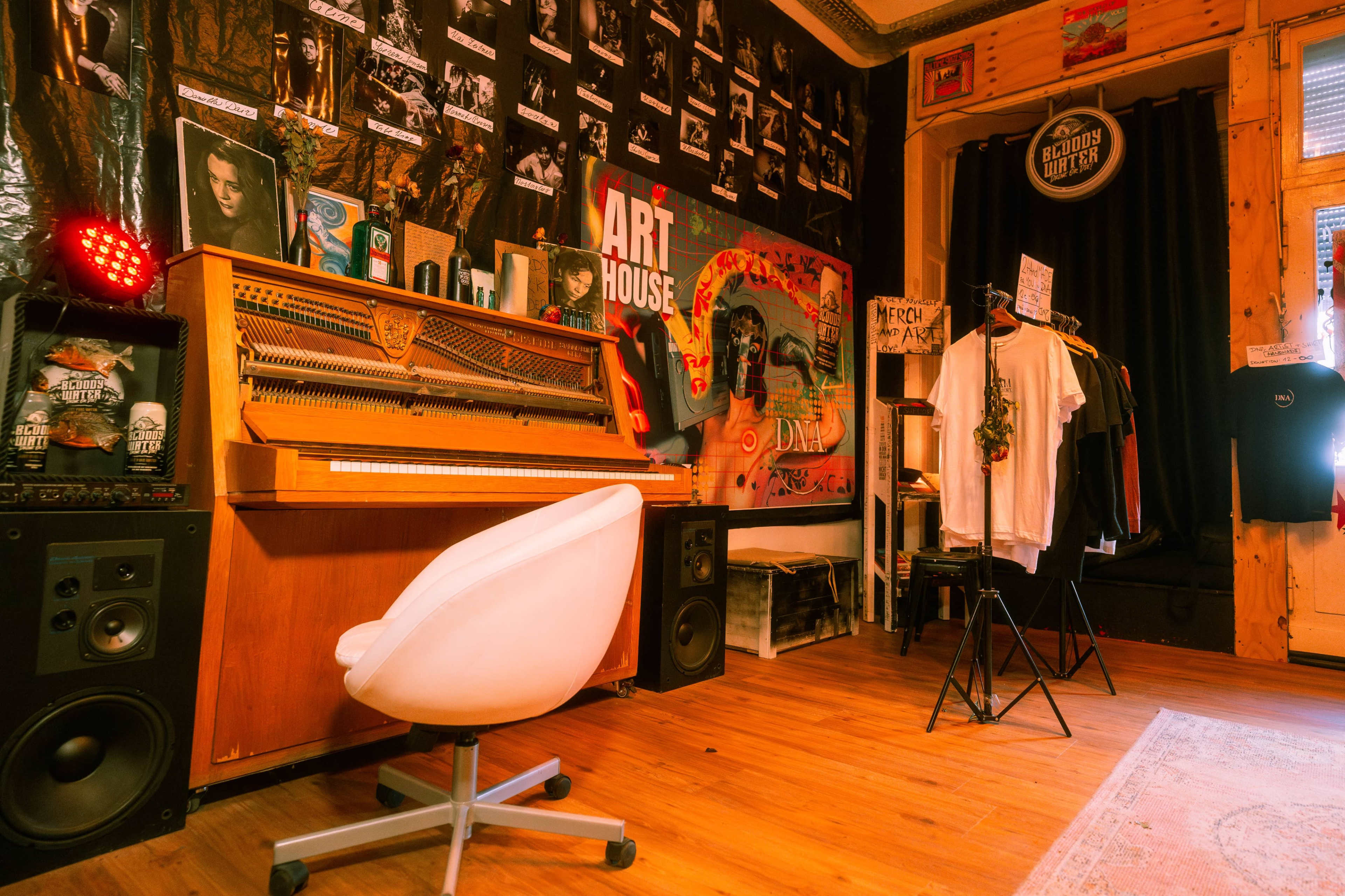 The image shows a stylish room featuring a wooden piano, a collection of posters on the walls, a clothing rack with shirts, and speakers positioned next to the piano.