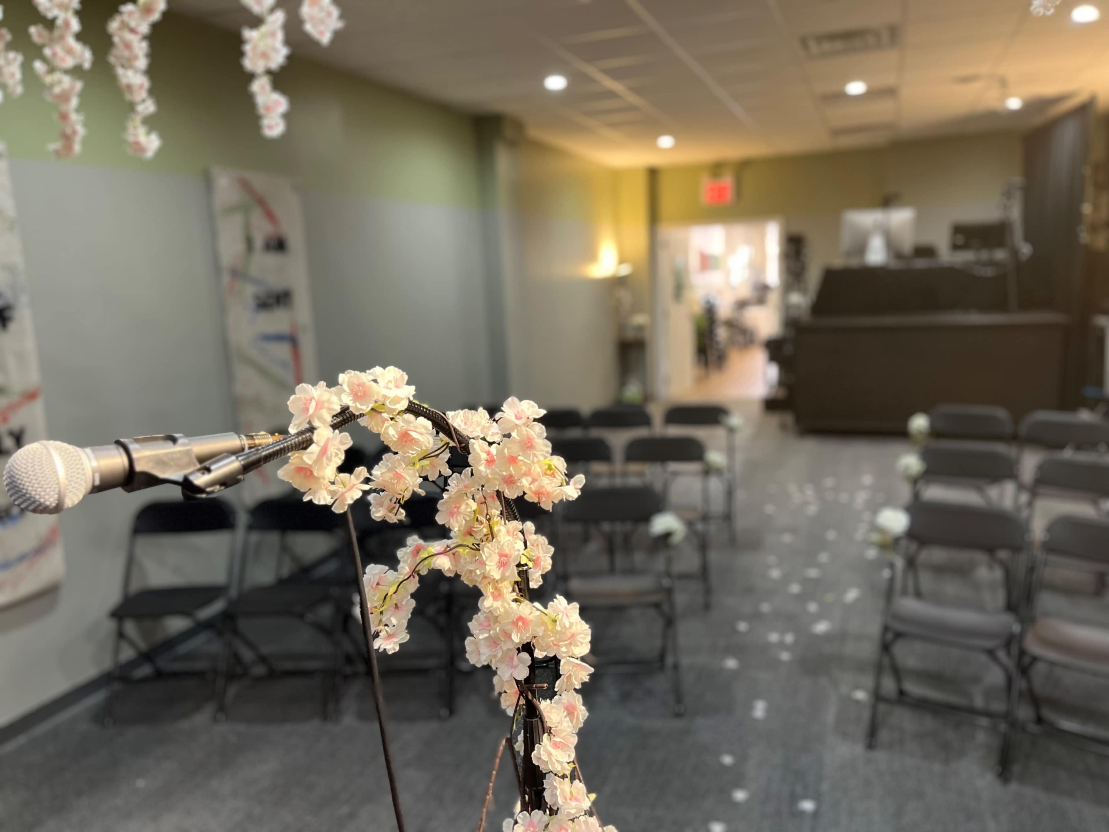 The image shows a microphone in the foreground with a floral decoration, and a room arranged with chairs for an event, leading to an open doorway in the background.