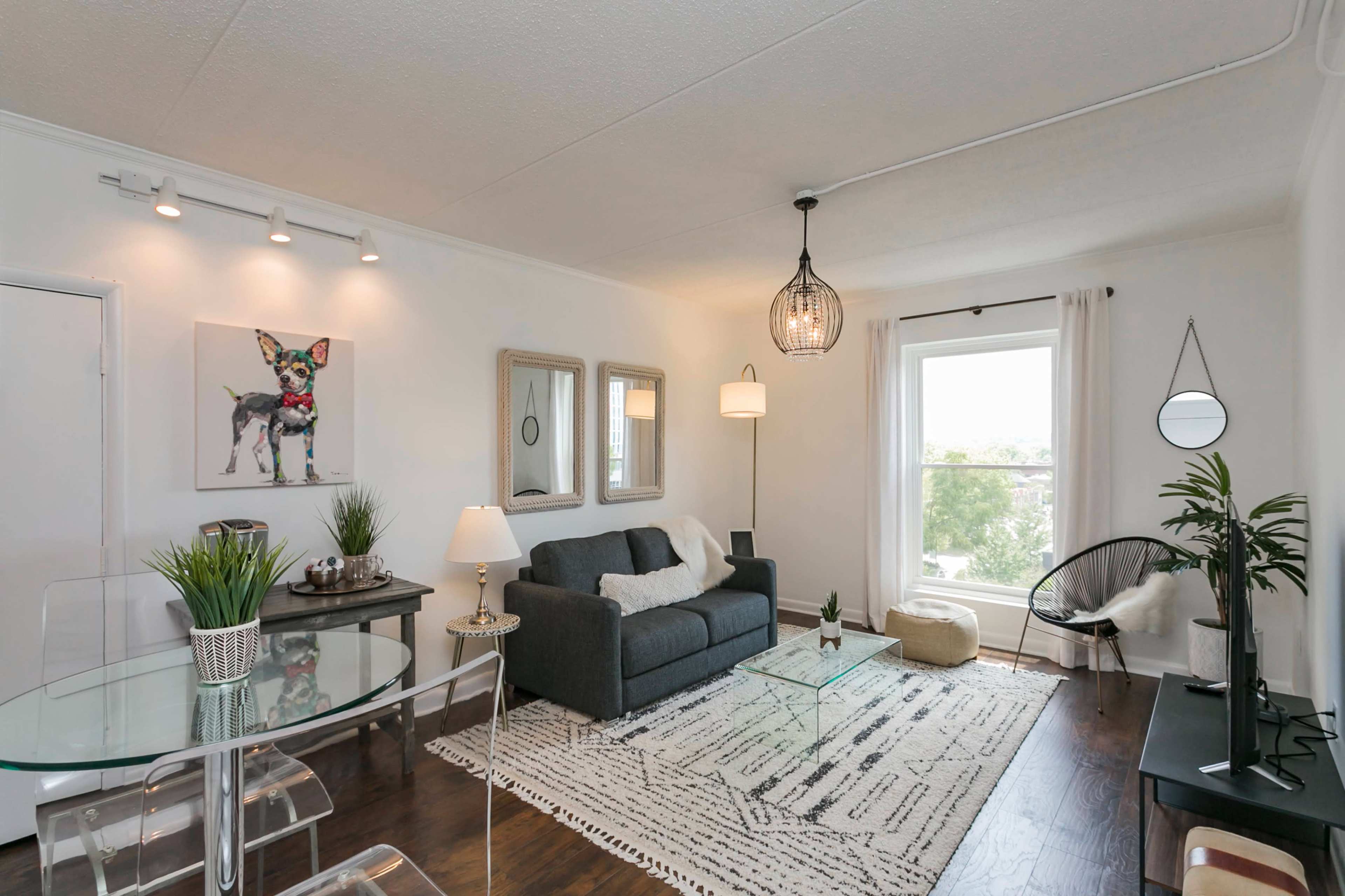 The image shows a modern living room featuring a gray sofa, a glass coffee table, and a large window, with decorative plants and artwork on the walls.