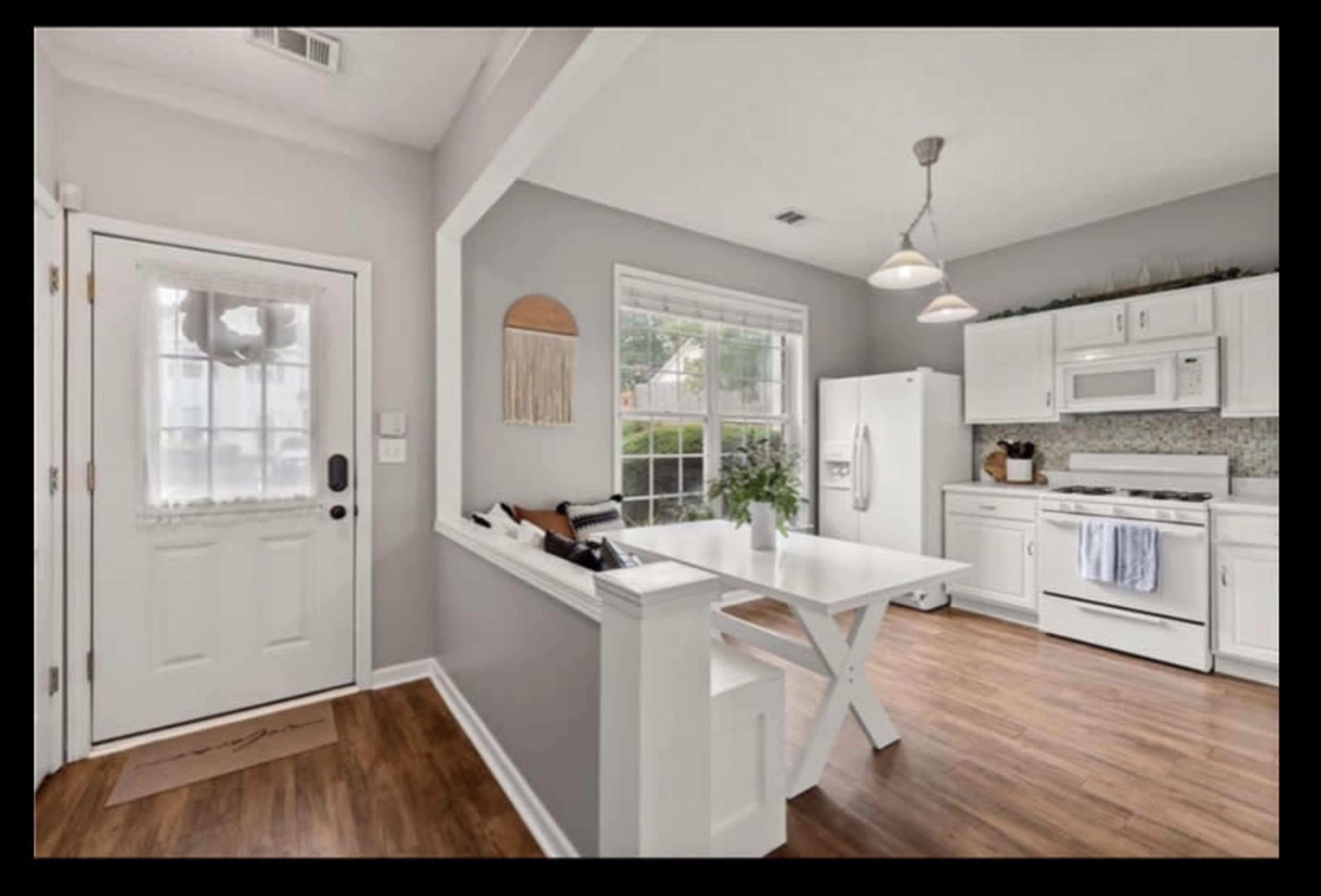 The image shows a modern kitchen and dining area with white cabinetry, a central table, and natural wood flooring.