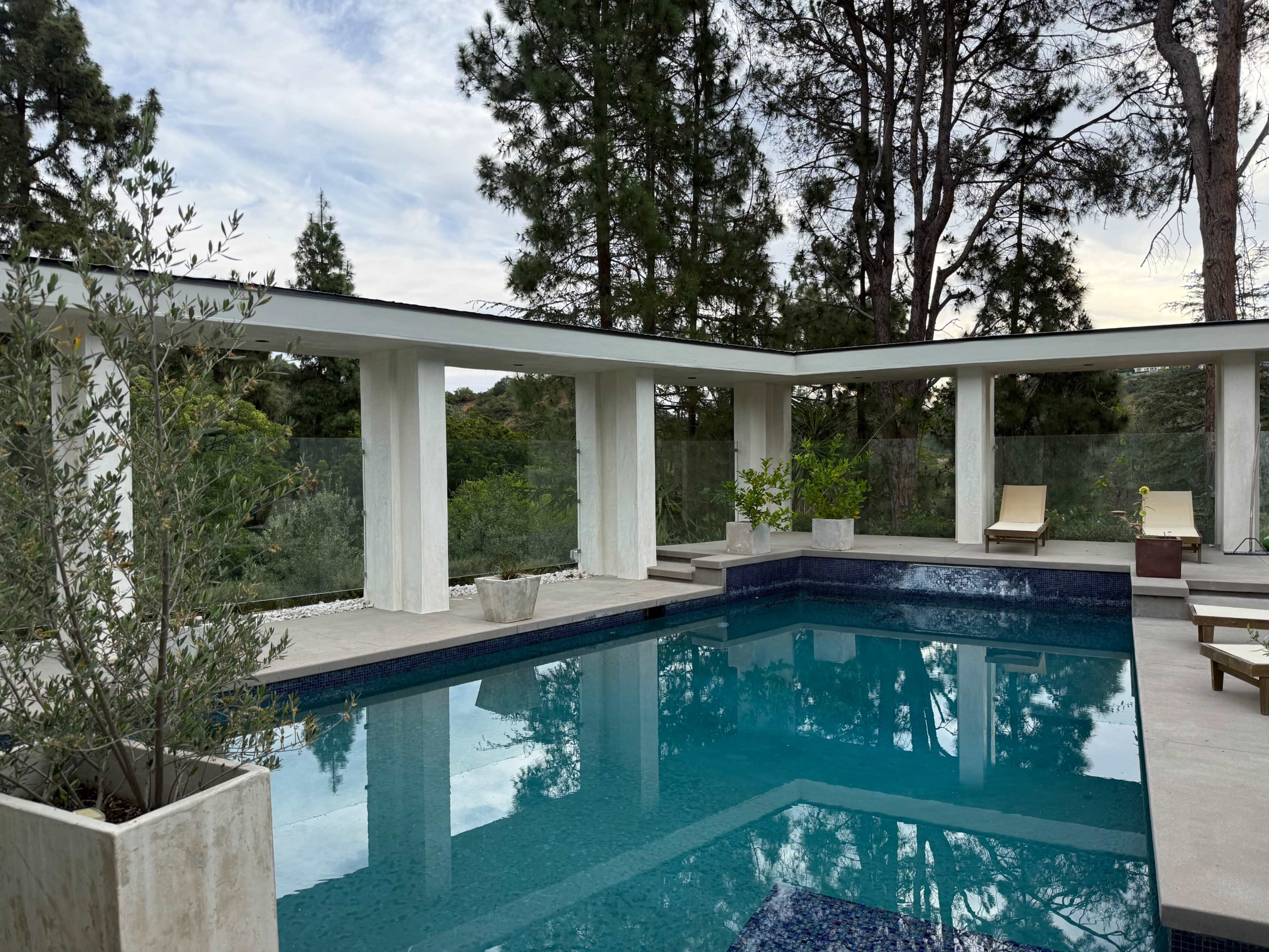 The image shows a modern pool area surrounded by trees, with lounge chairs on a concrete deck and large glass windows providing a view of the landscape.