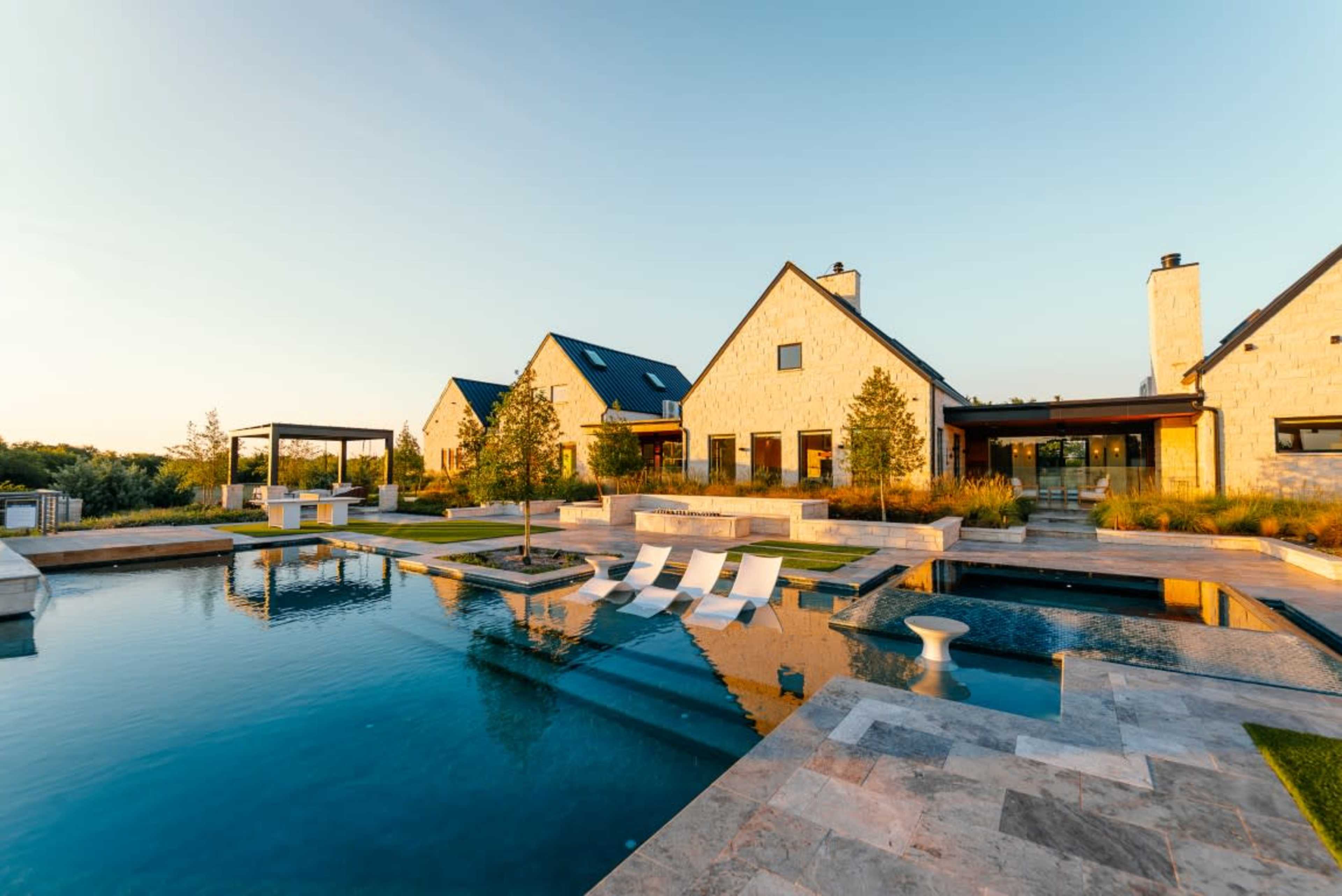 The image shows a modern house with stone facades overlooking a rectangular swimming pool, surrounded by landscaped gardens and outdoor seating areas.