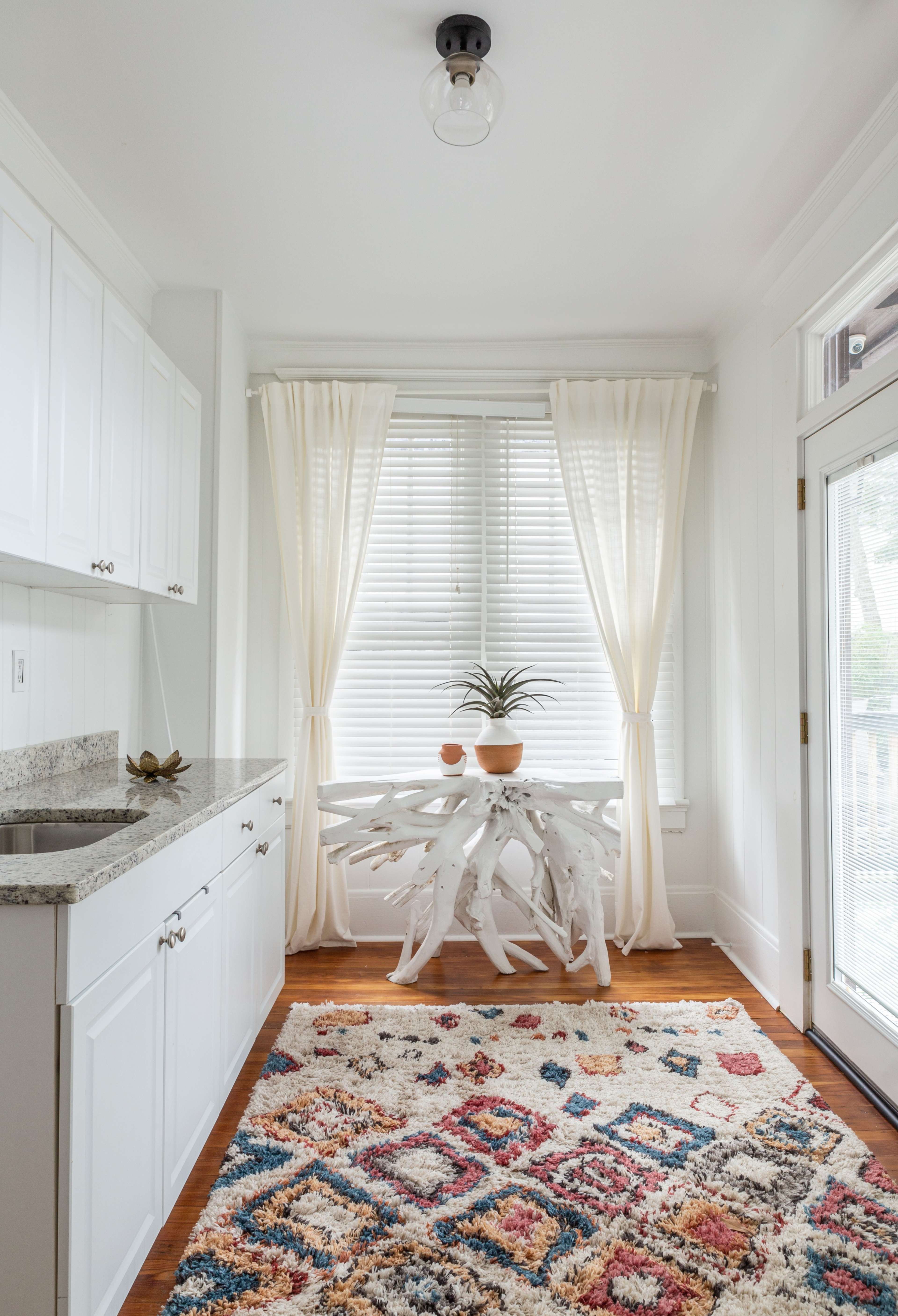 A sunlit room features white cabinetry, a decorative table with a plant, and a colorful patterned rug on the wooden floor.