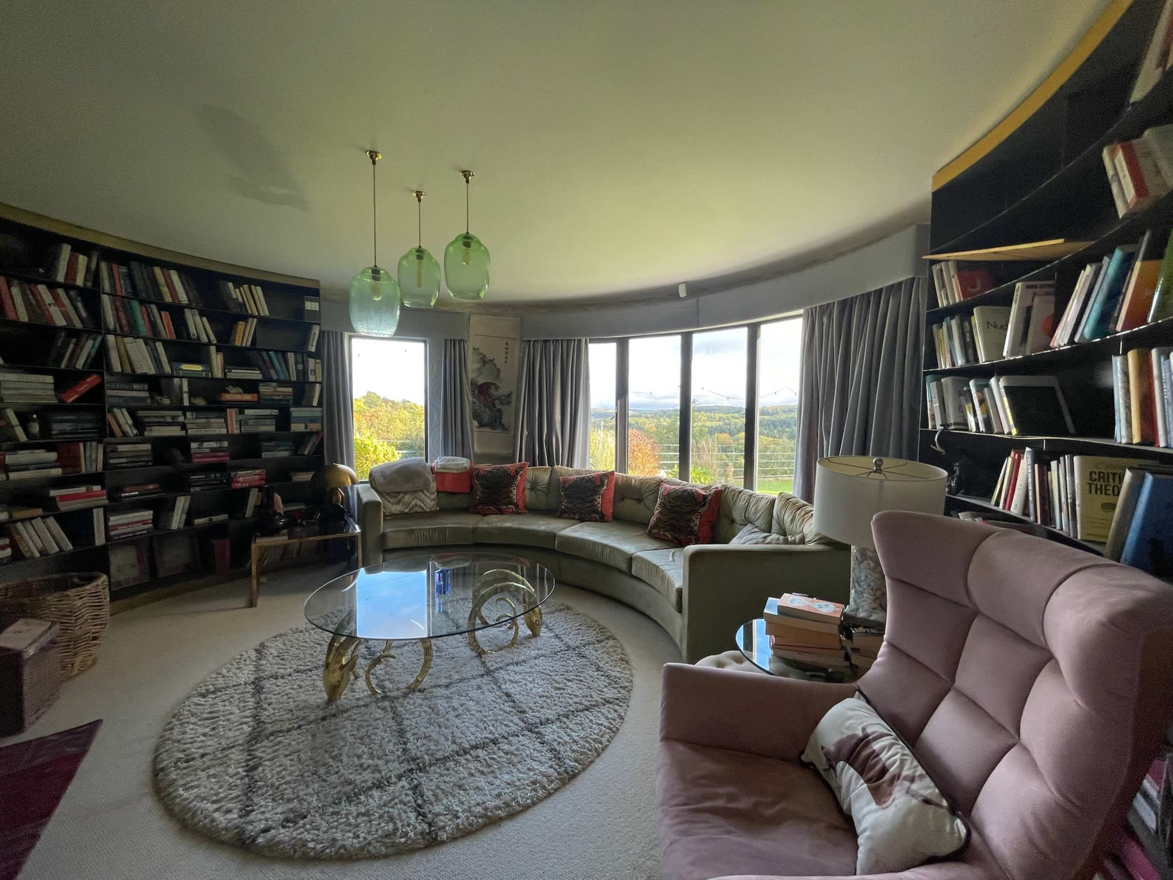 The image shows a cozy reading area with a curved sofa, a round glass coffee table, and floor-to-ceiling bookshelves filled with books, overlooking a scenic view through large windows.