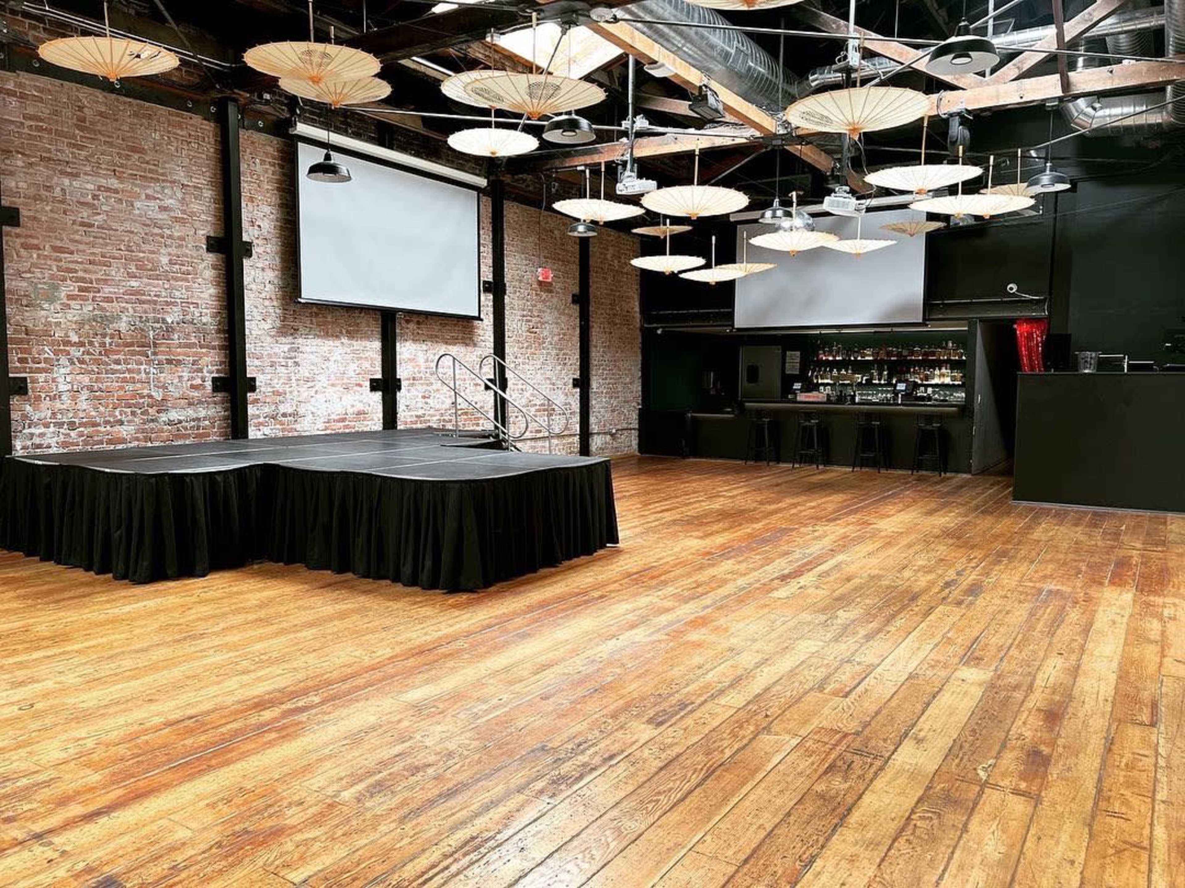 The image shows a spacious event venue with exposed brick walls, a wooden floor, a small stage, and two large screens.