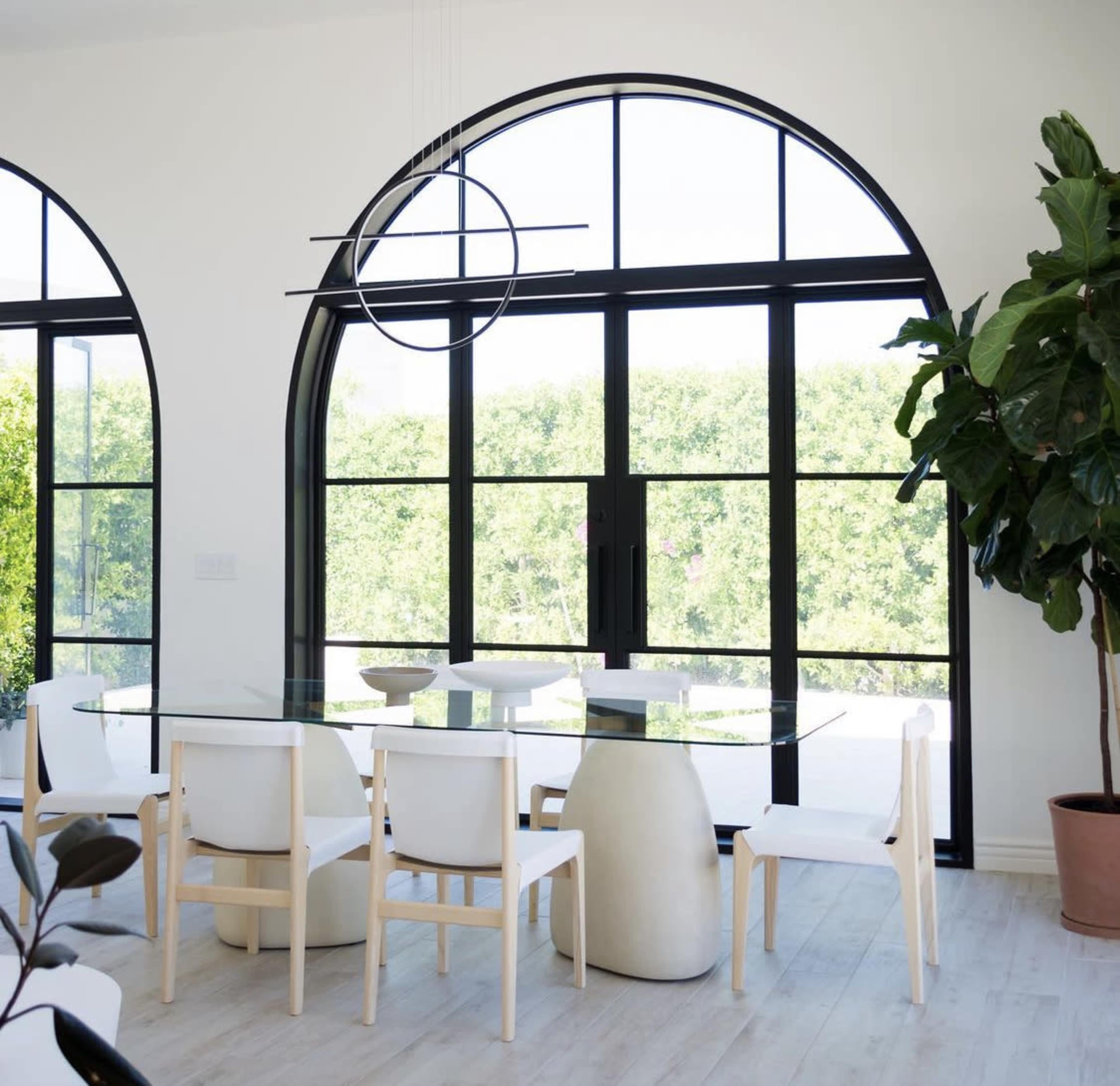 A modern dining area features a glass table surrounded by white chairs, with large arched windows offering a view of greenery outside.