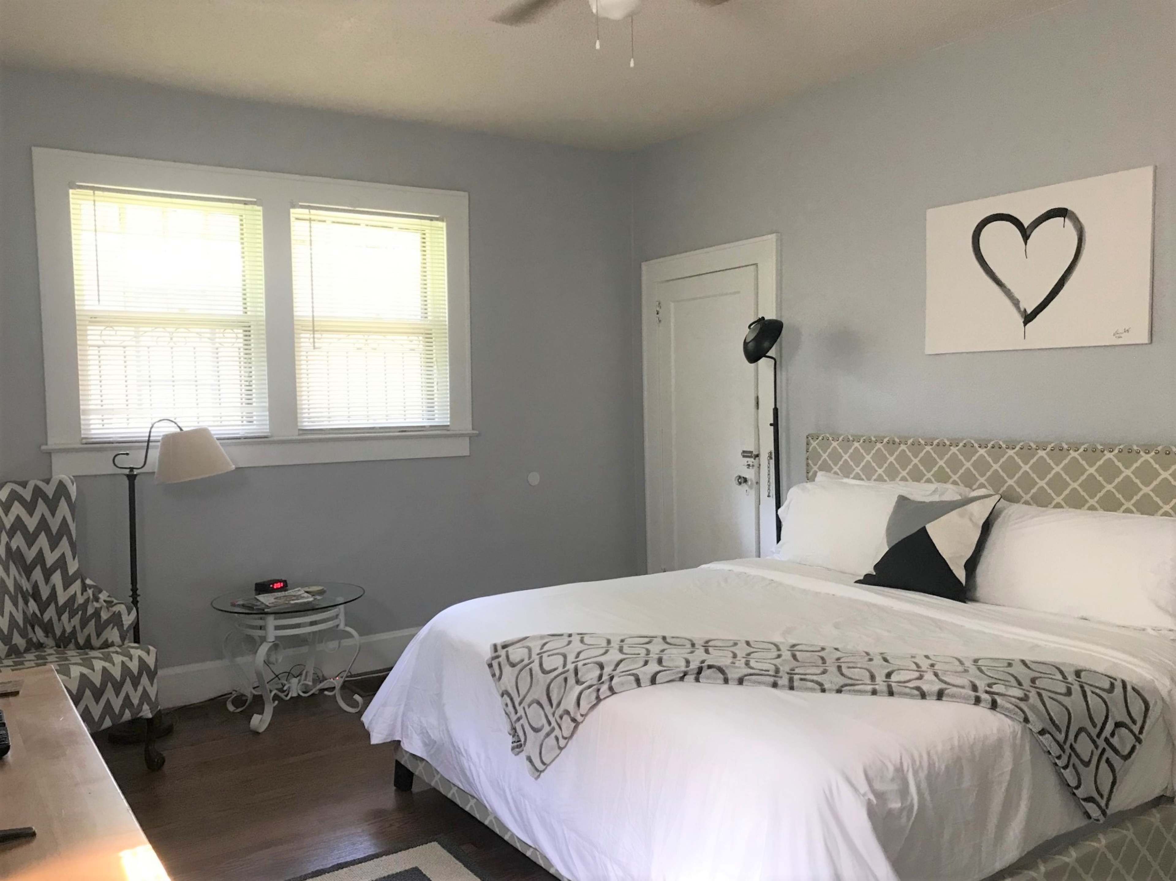 The image shows a bedroom with a bed, a chair, a small side table, and a piece of artwork featuring a heart on the wall.