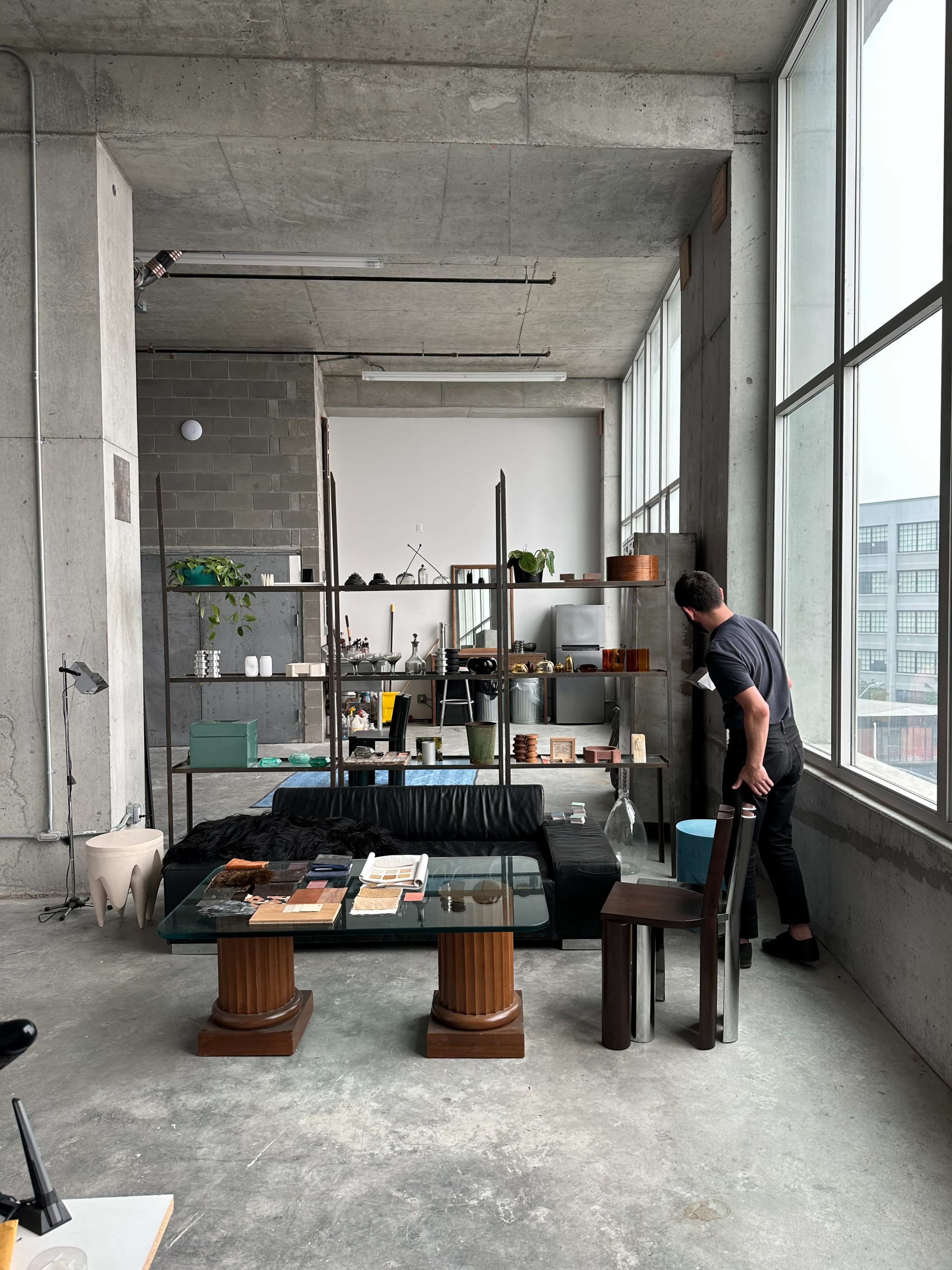 A man stands beside a modern shelf filled with decorative items in a minimalist industrial-style living space with large windows.