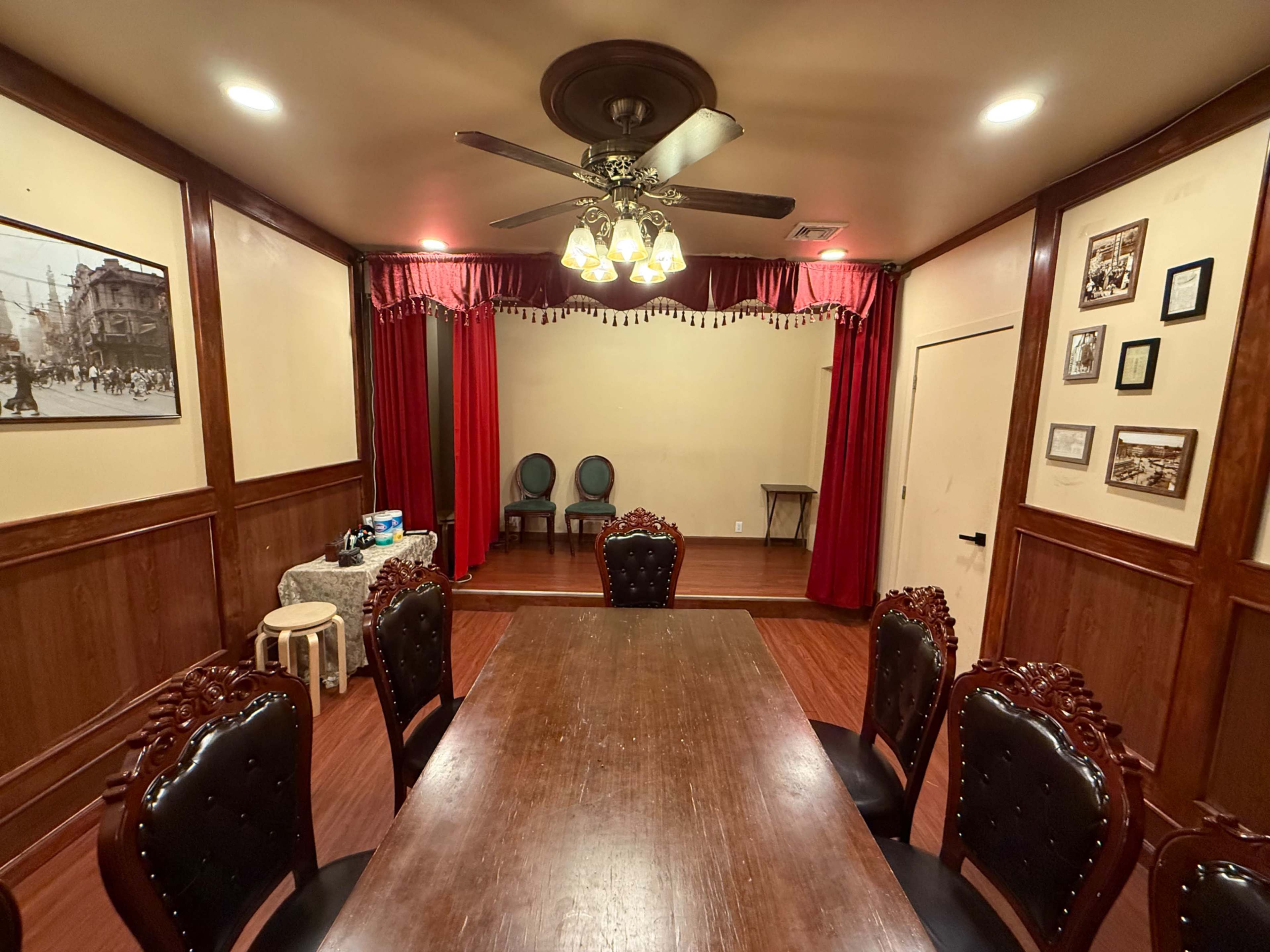 The image shows a banquet-style room with a large wooden table and ornate chairs, featuring a stage with red curtains at one end.