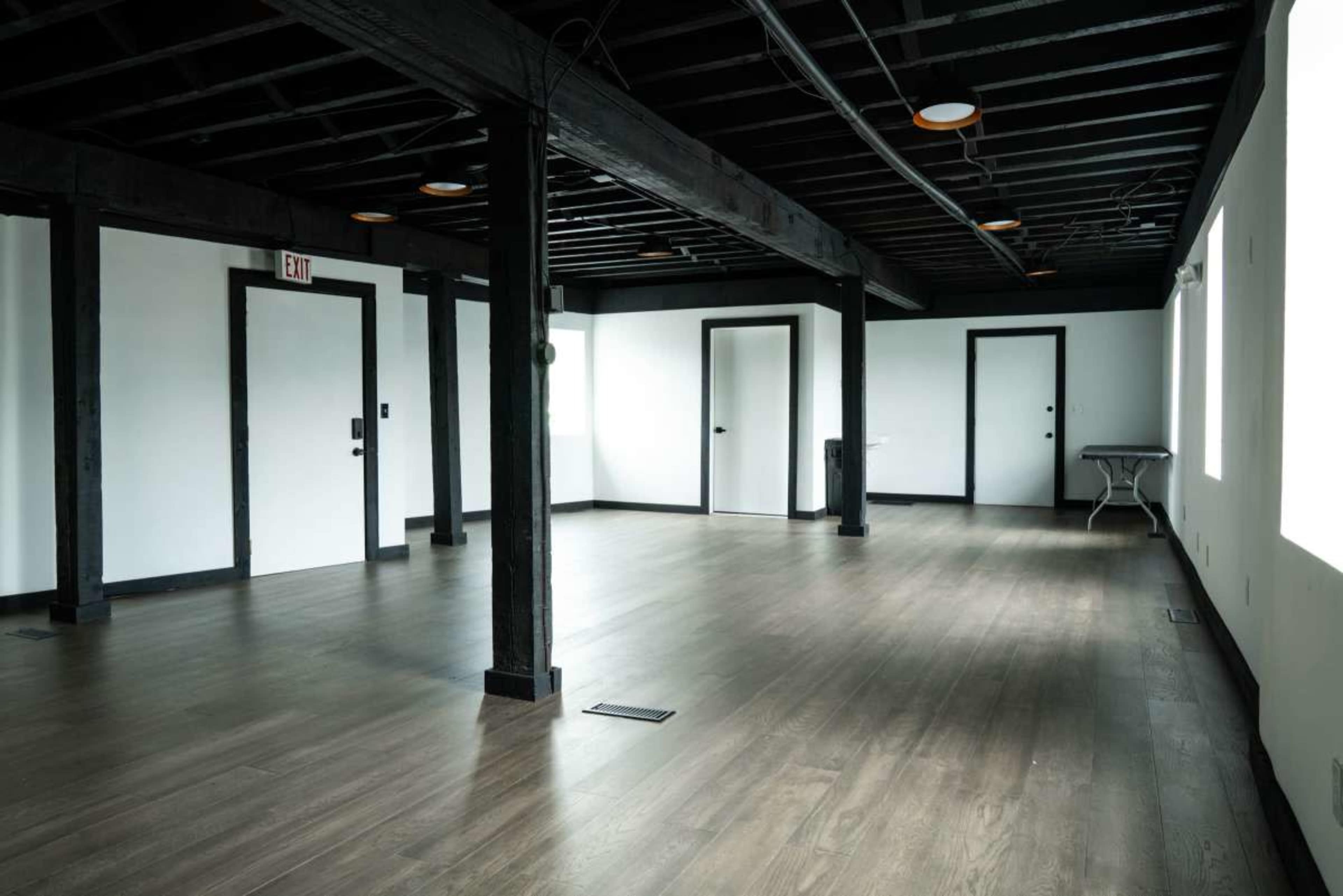 The image shows an empty room with wooden beams and several doors, featuring a table against one wall and large windows allowing natural light to enter.