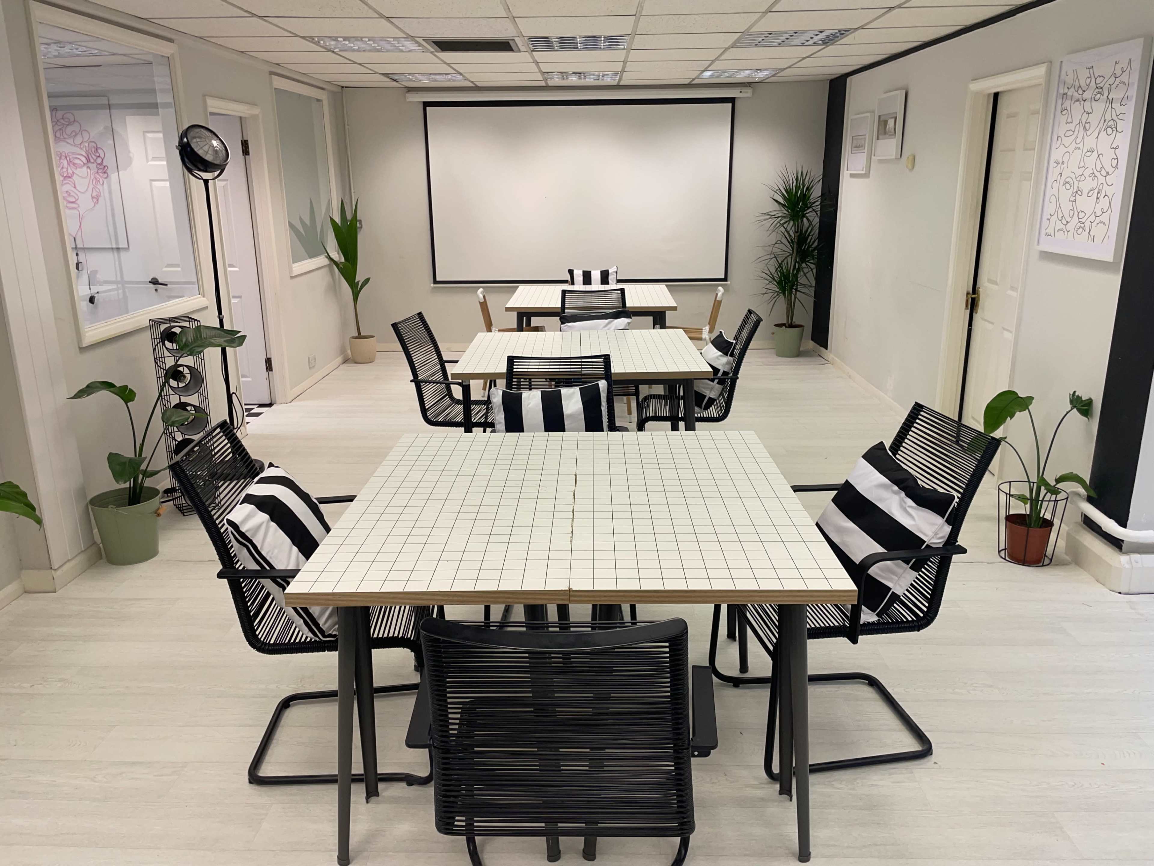 A modern meeting room features several square tables with striped chairs, surrounded by potted plants and a projector screen at the front.