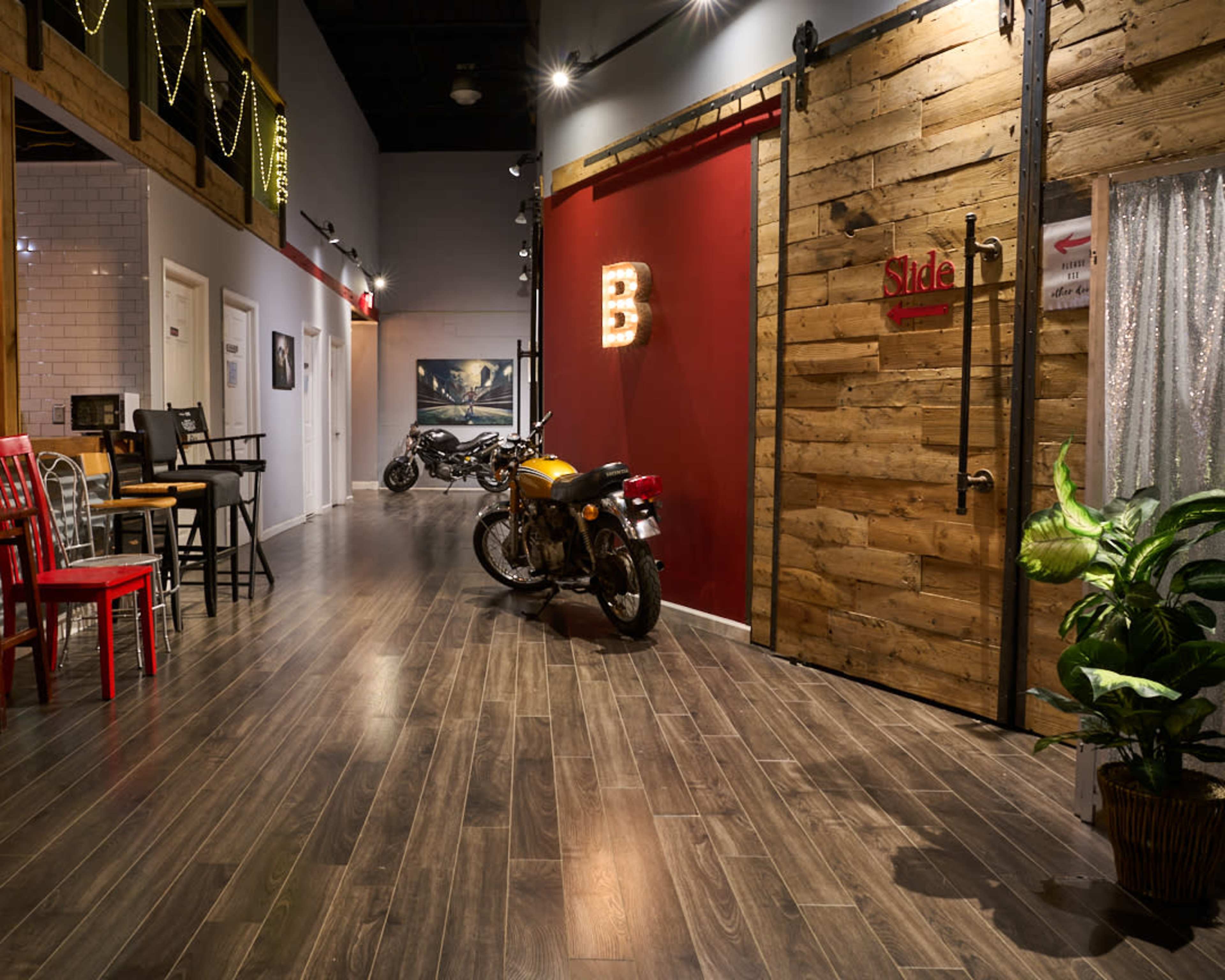 The image features a modern hallway with a wooden door, a motorcycle, colorful chairs, and dim lighting accentuating the décor.