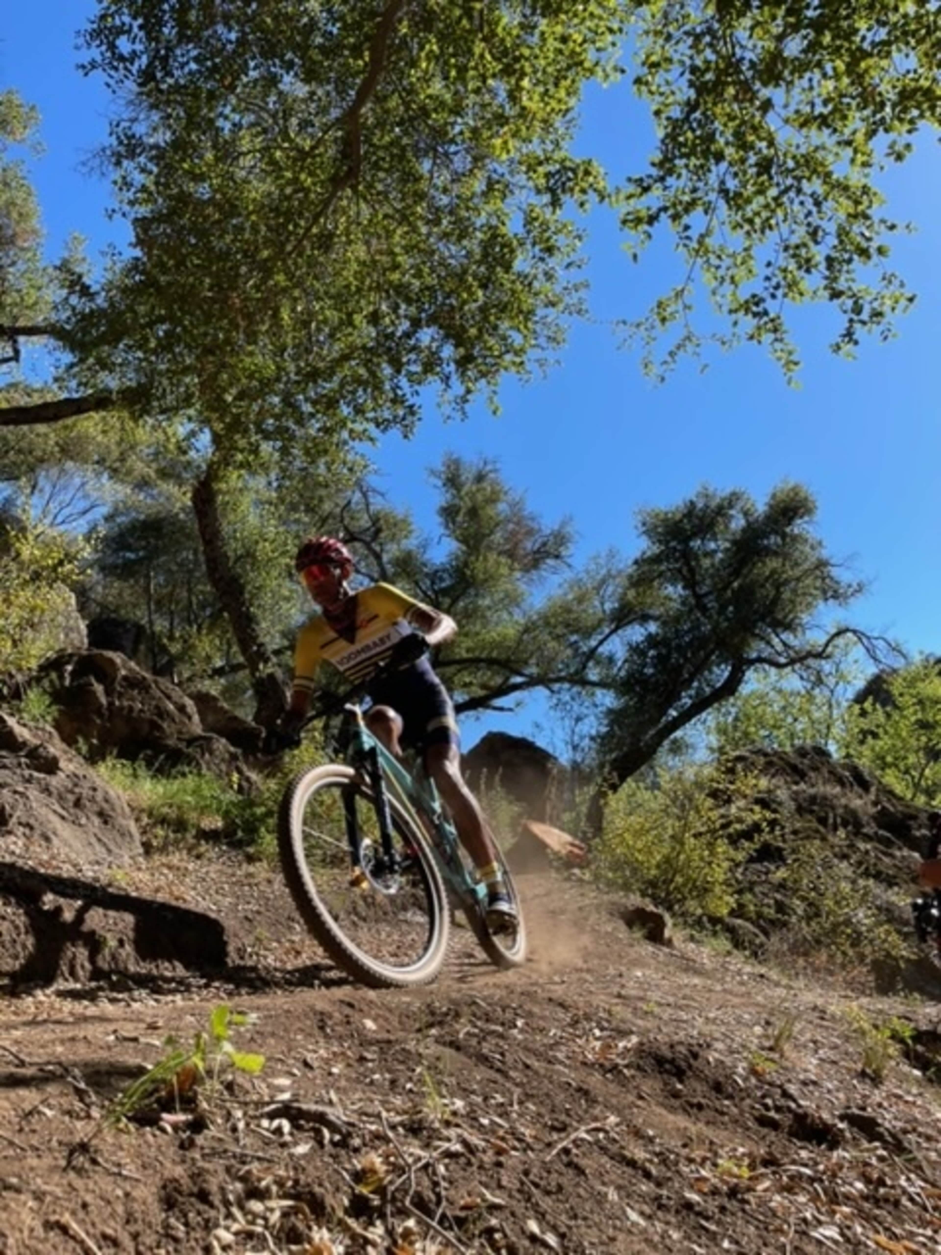 A cyclist rides a mountain bike along a dirt trail surrounded by trees and rocky terrain.