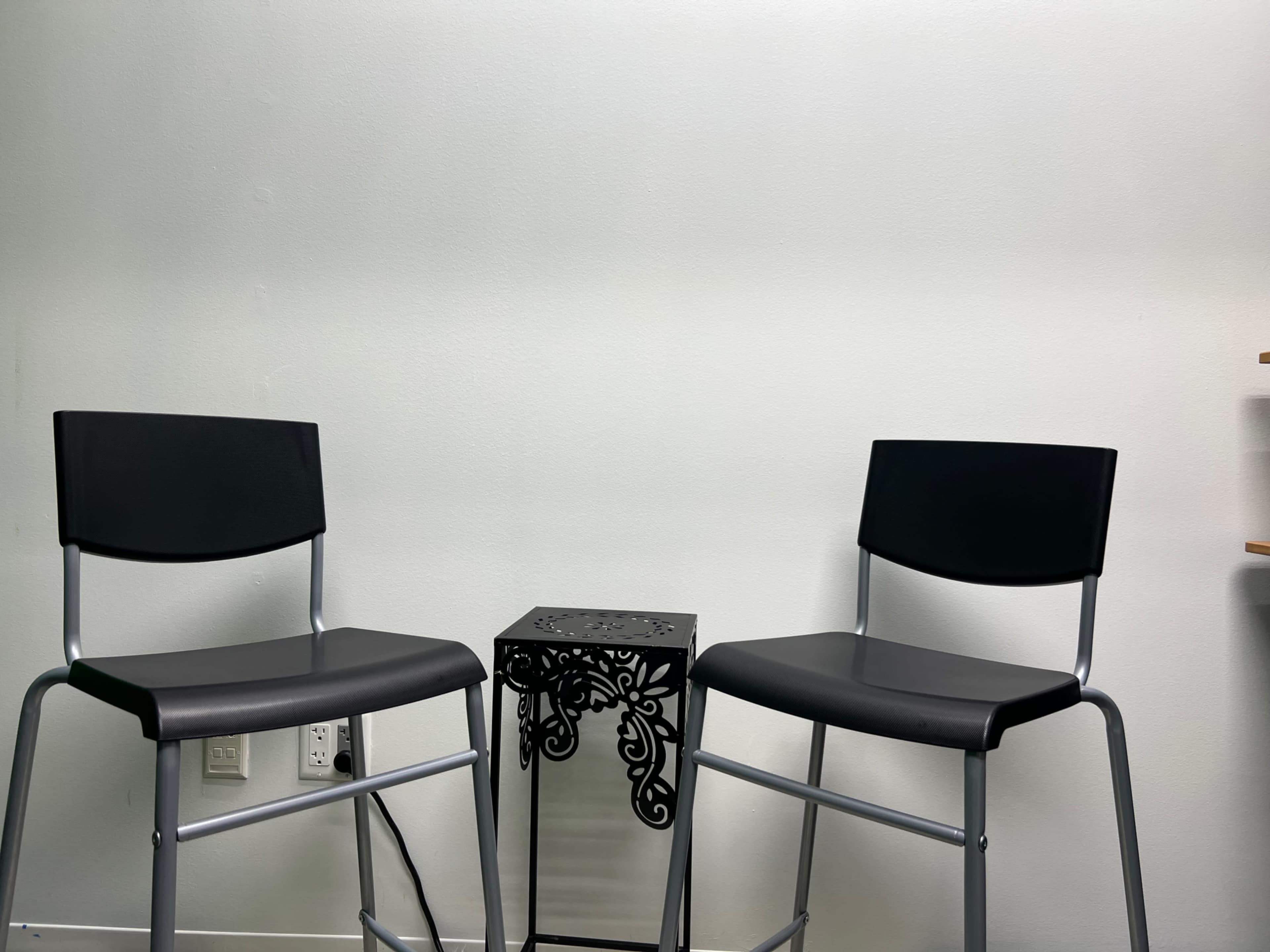 Two black stools are positioned on either side of a small, ornate metal table against a plain white wall.