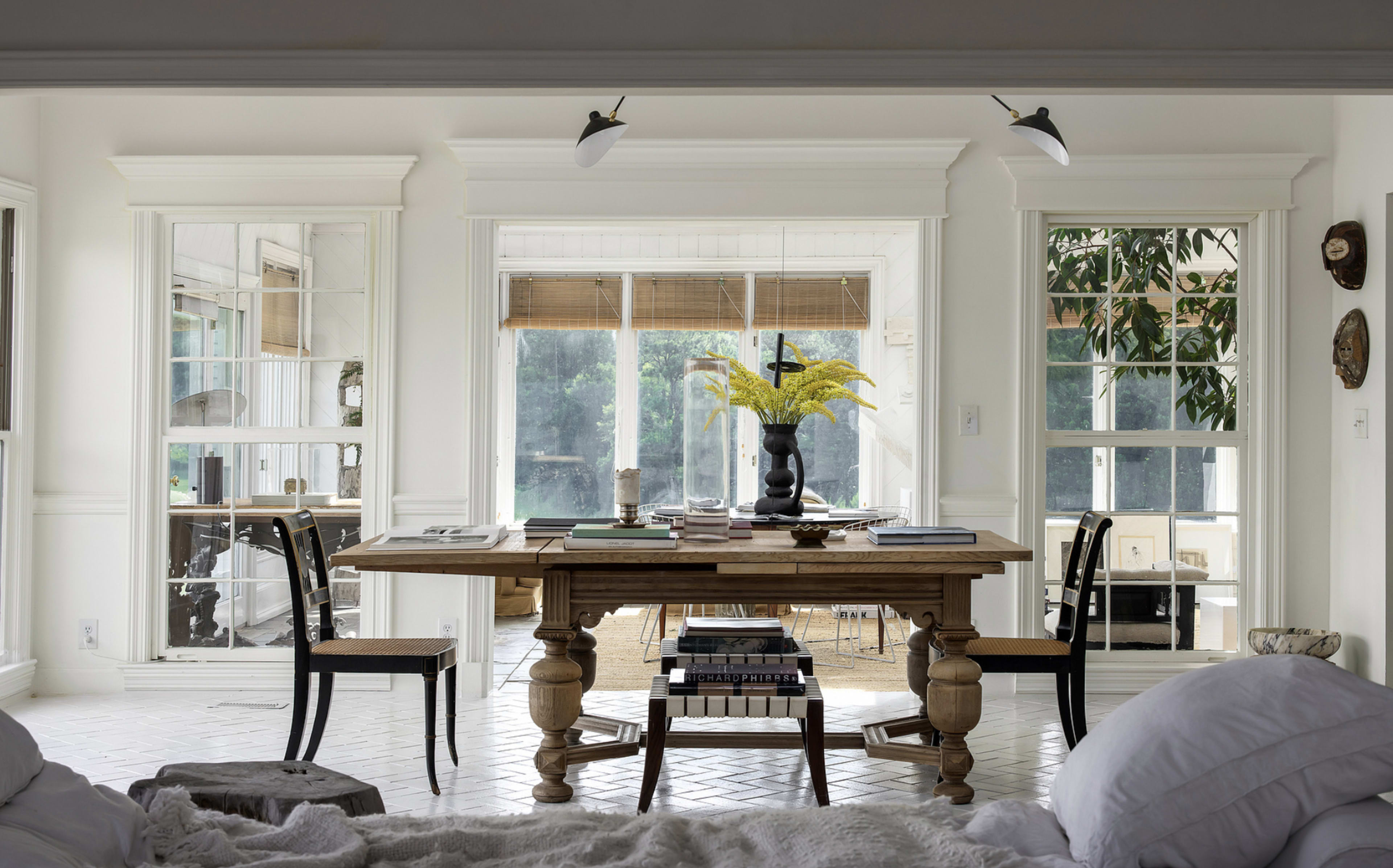 A wooden dining table is positioned in a bright room with large windows that overlook a green outdoor space, flanked by two black chairs.