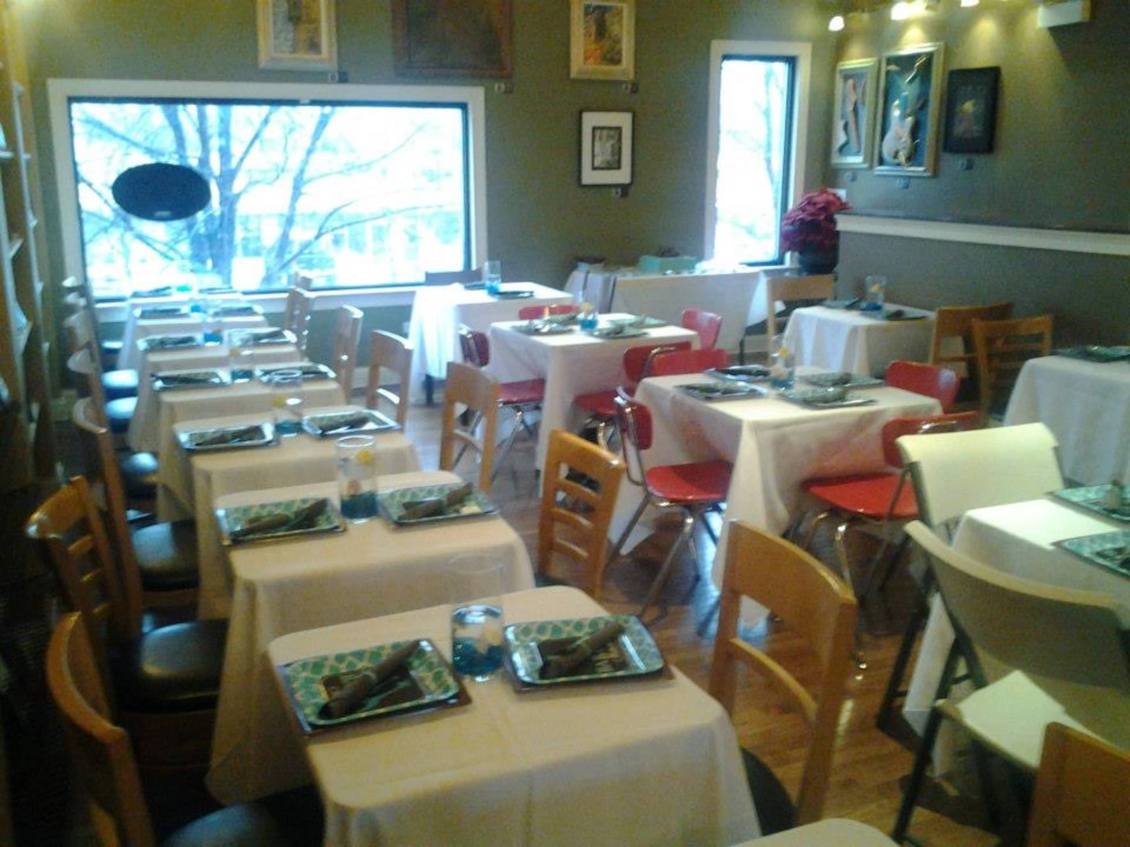 A dining area with neatly set tables covered in white tablecloths, arranged in rows, with chairs of various colors surrounding them.