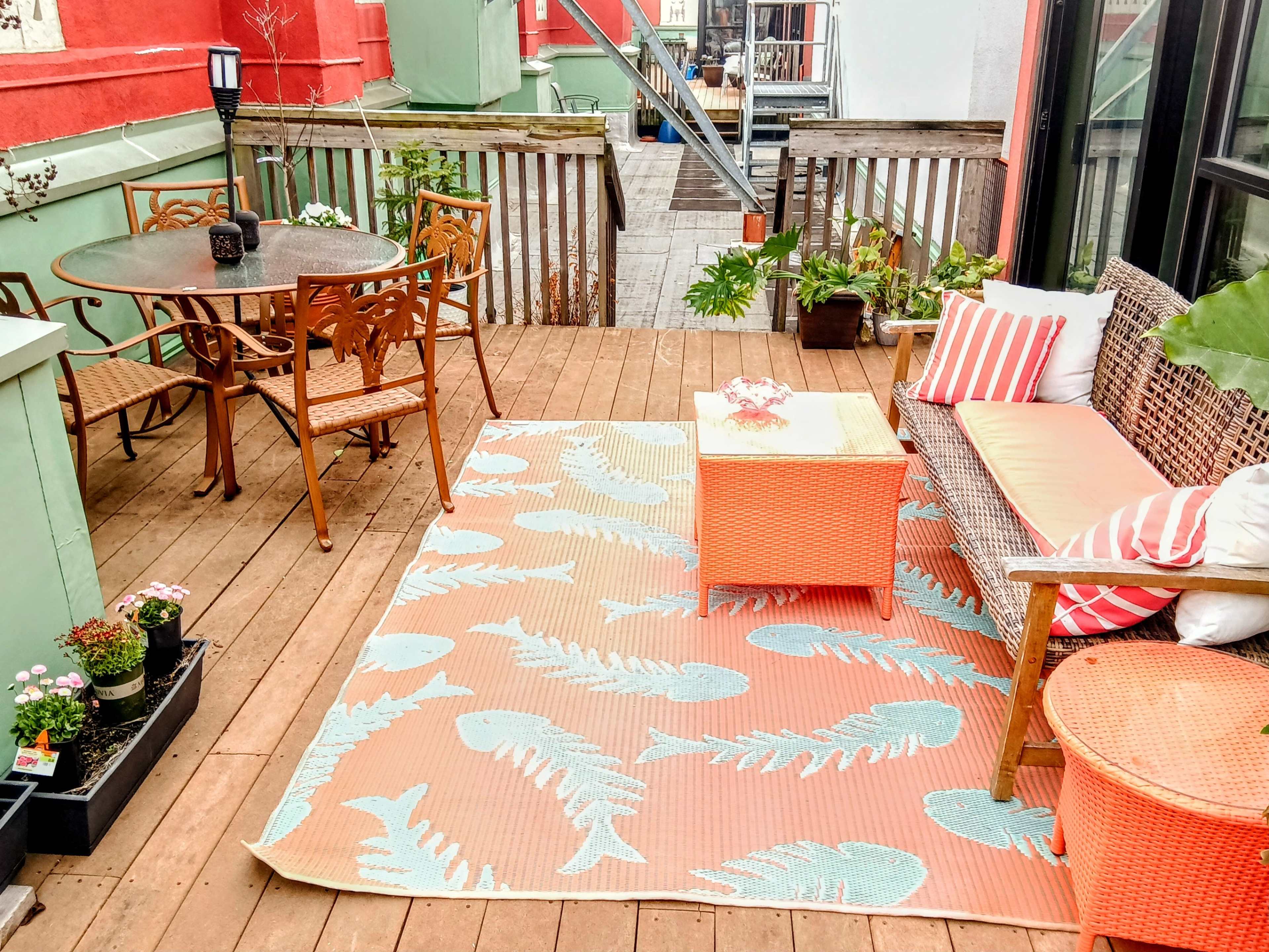 A rooftop terrace features a wooden deck, a patterned rug, outdoor furniture, and potted plants.