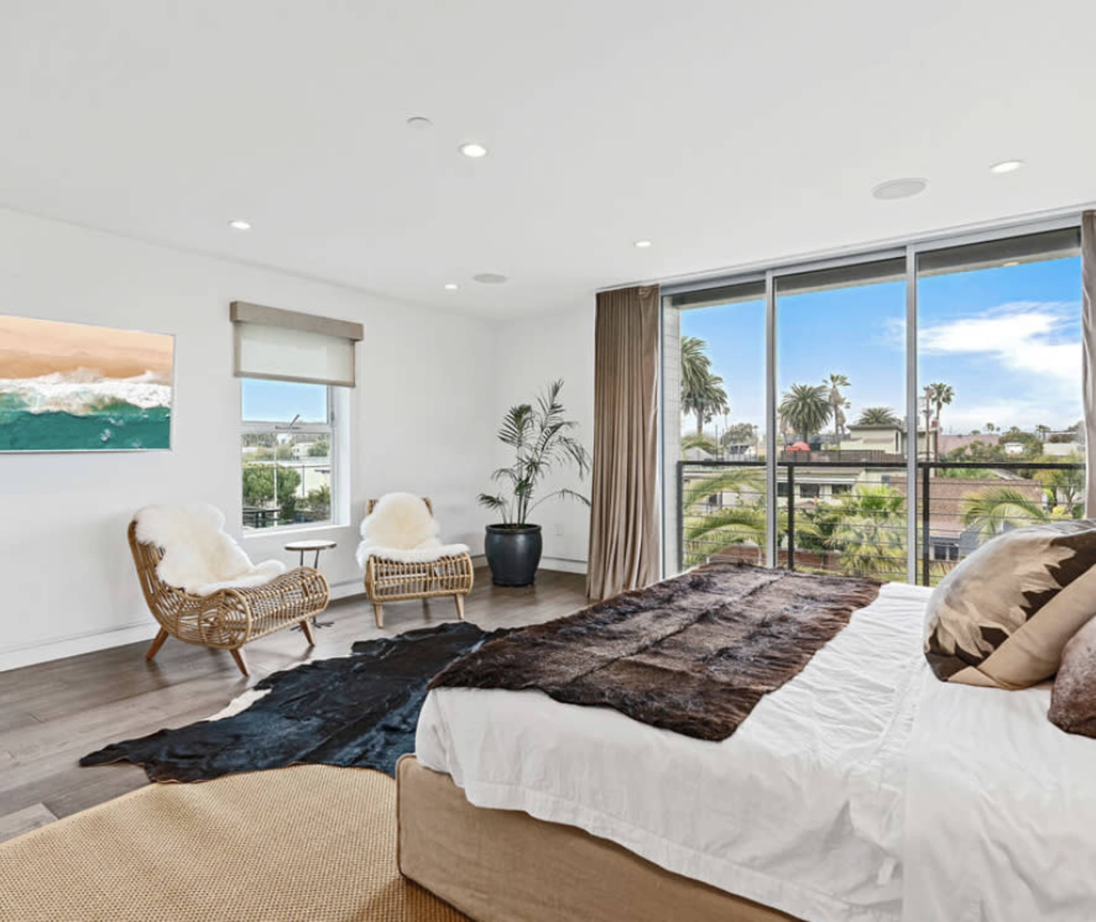 The bedroom features a large bed with a brown throw, two rattan chairs, a potted plant, and large windows showcasing a view of palm trees and a clear sky.