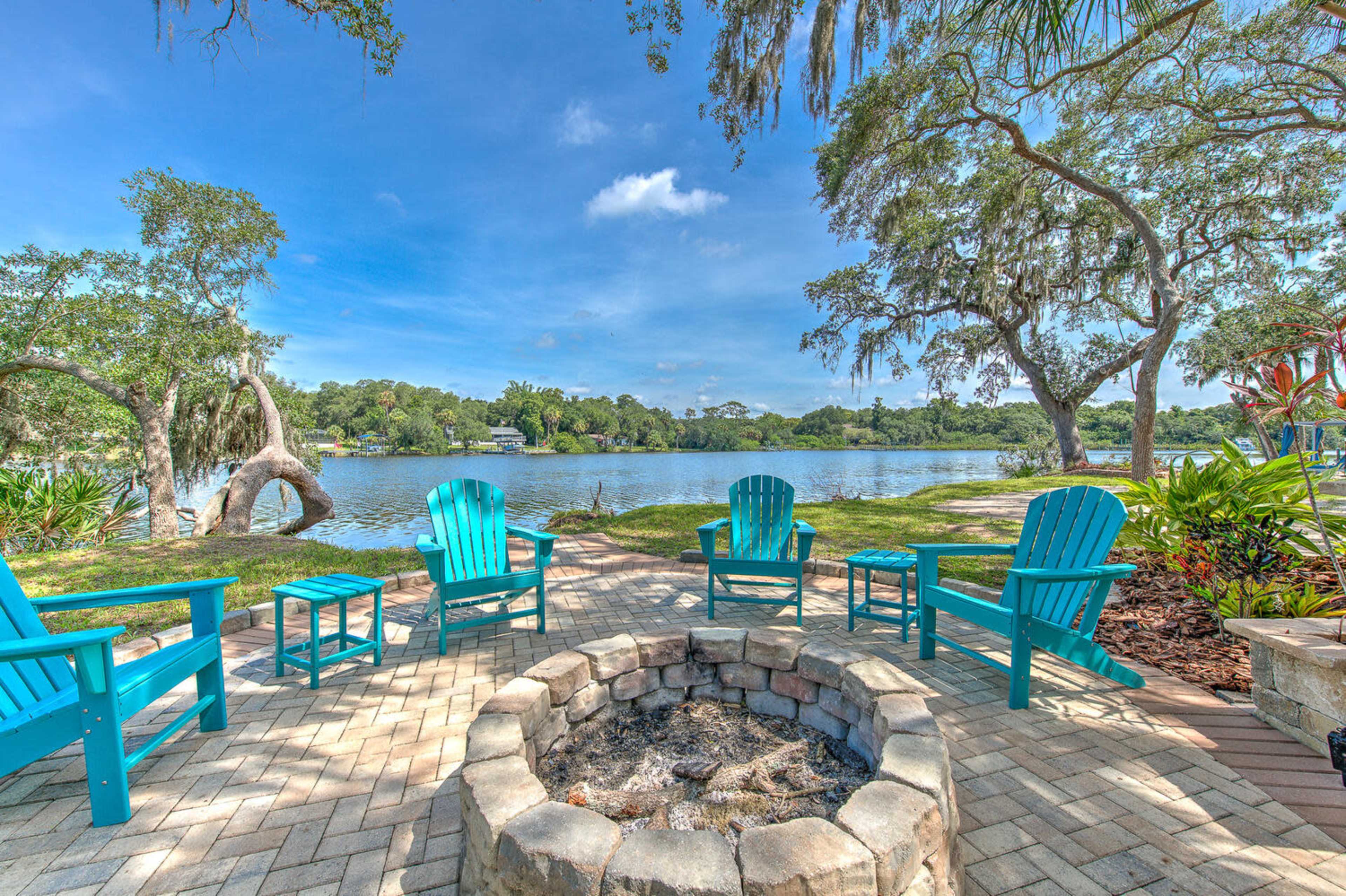 A seating area with turquoise chairs surrounds a stone fire pit next to a calm lake, framed by trees and greenery.
