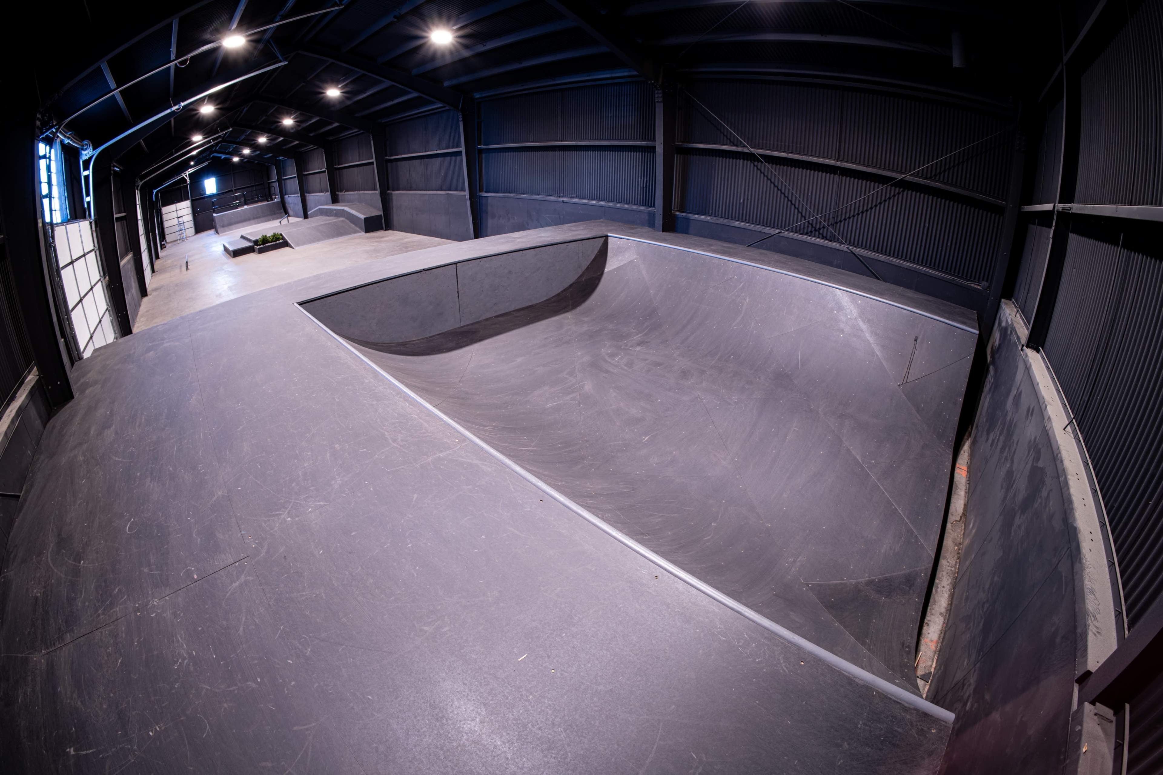 The image shows a spacious indoor skatepark featuring a large, empty bowl-shaped ramp and several other skateboarding surfaces.