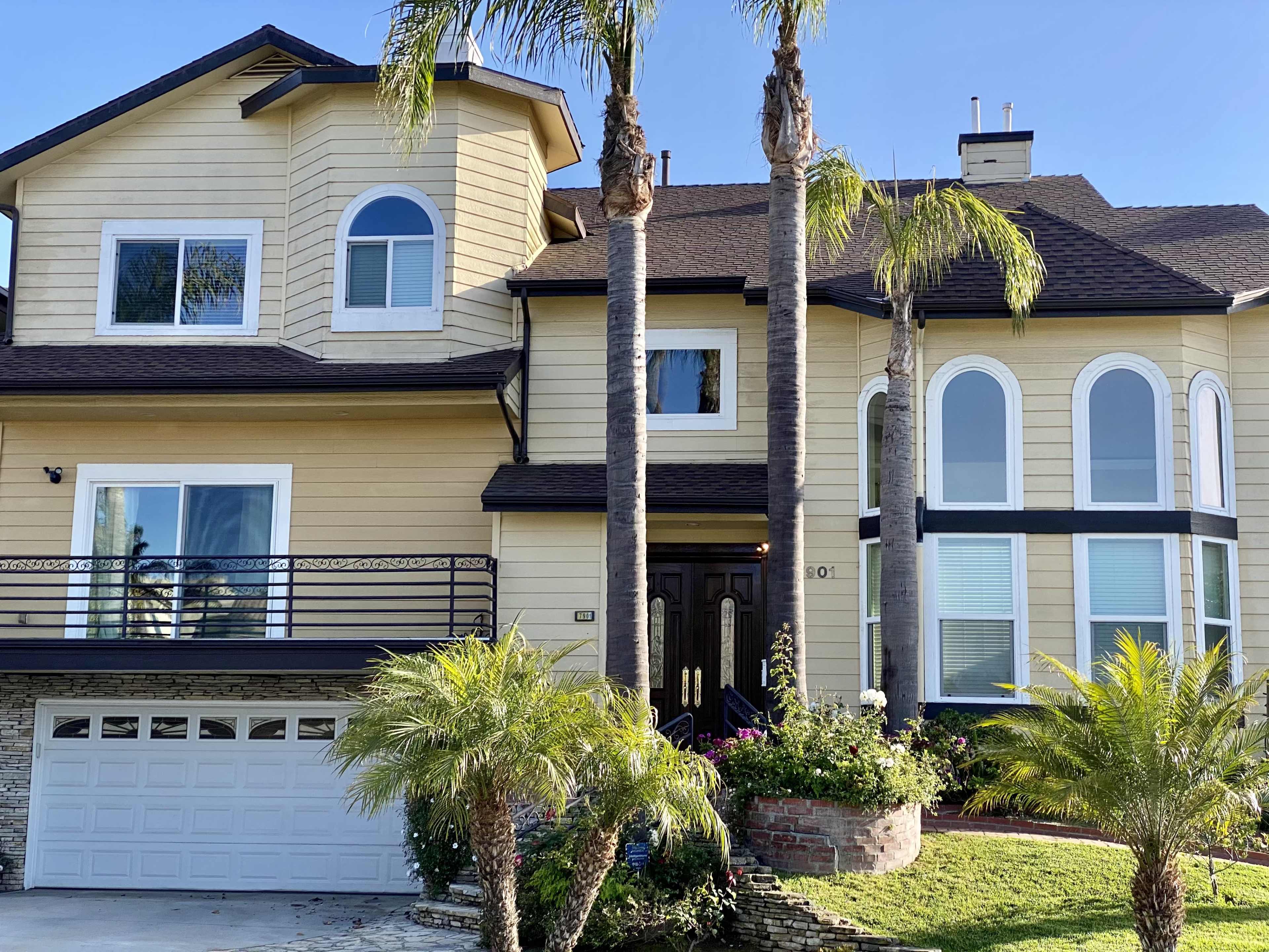 A large two-story house with a beige exterior, two palm trees in front, and a driveway leading to a garage door.