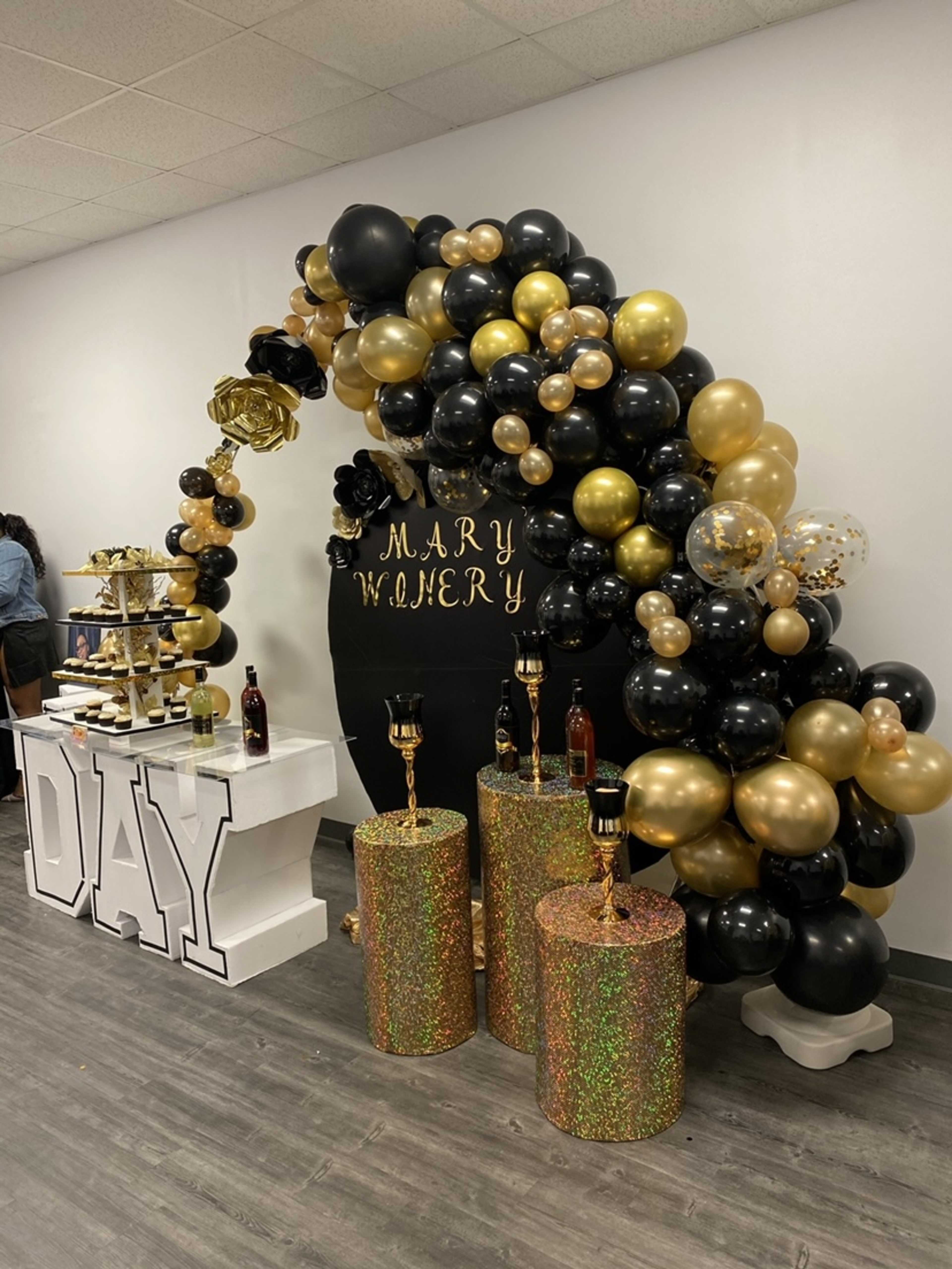 A festive decoration with a black and gold balloon arch, a display table for desserts, and elegant glittery pedestal stands at a celebration event.