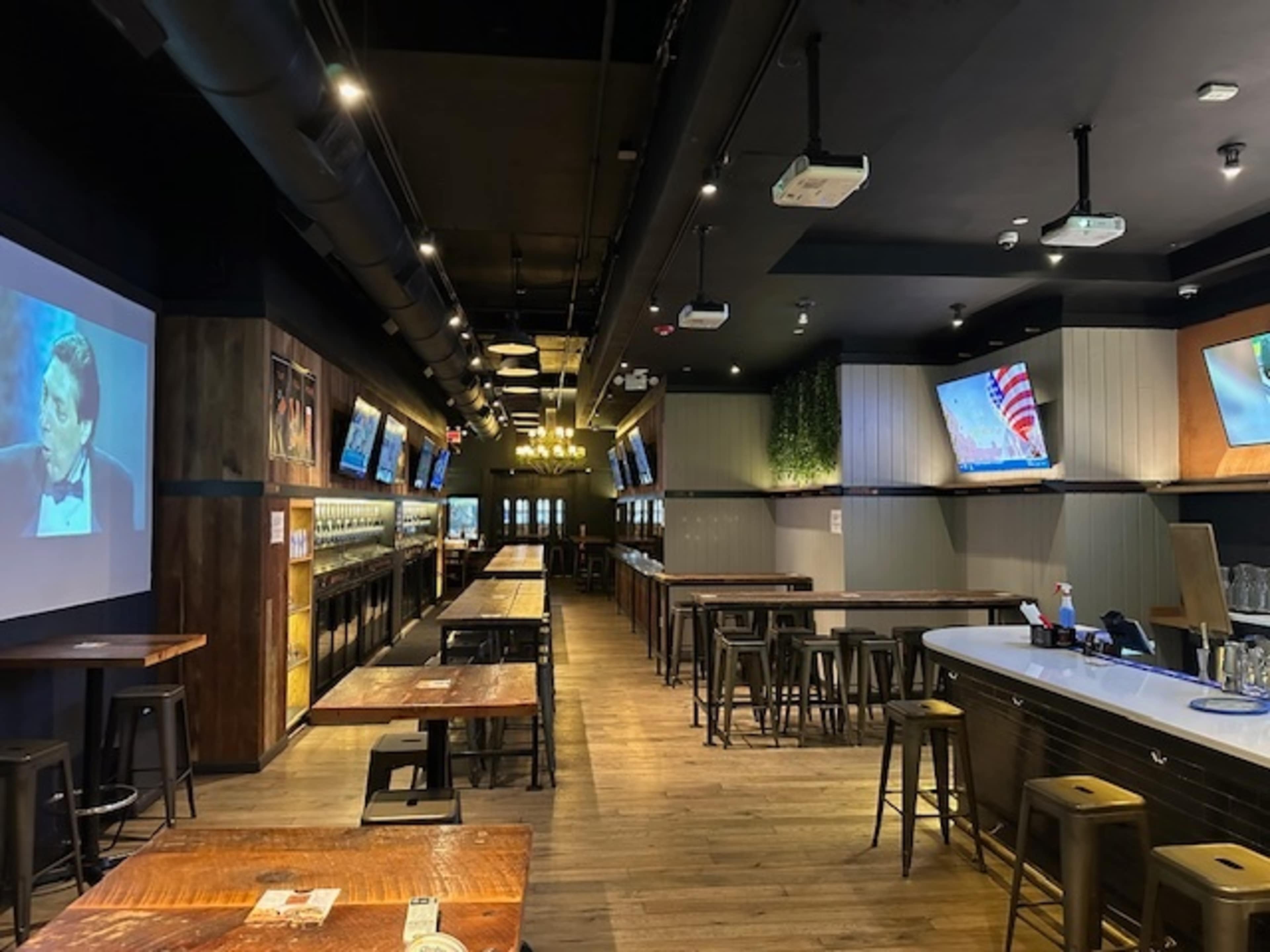 The image shows a modern bar interior with wooden tables, high stools, and several screens displaying sports broadcasts along the walls.