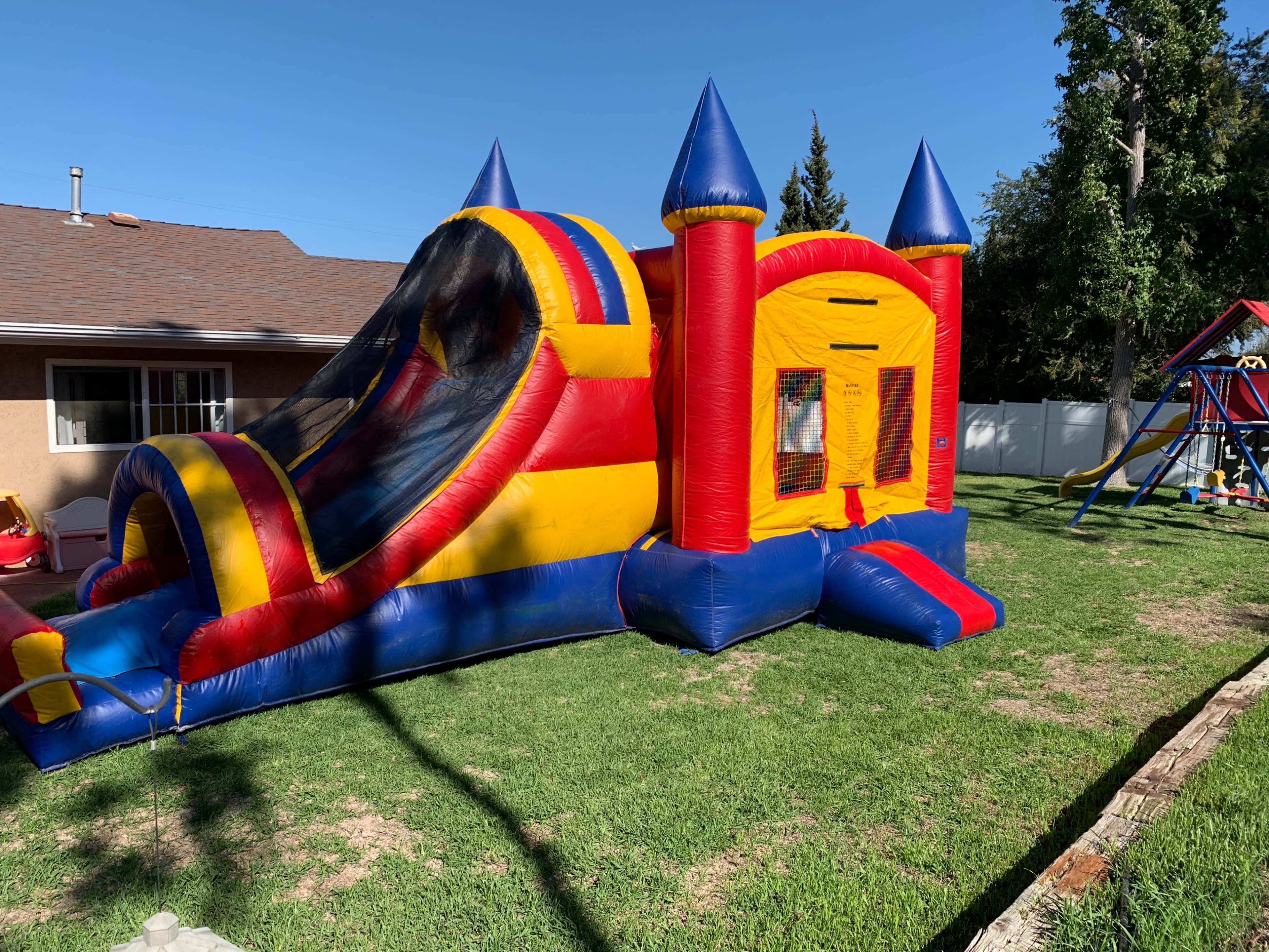 A large inflatable bounce house with a slide is set up on a grassy area next to a house.
