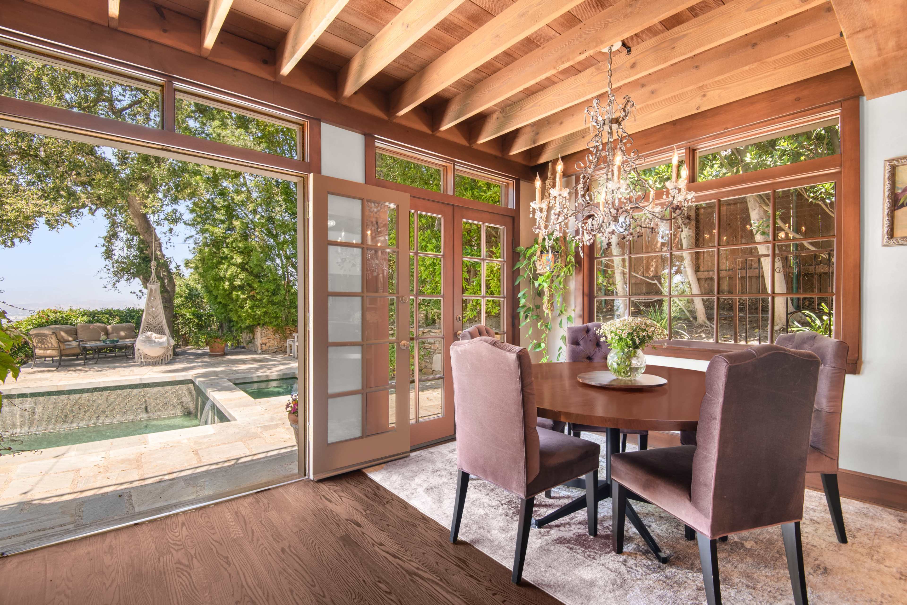 A dining area features a round table surrounded by upholstered chairs, with large windows overlooking a pool and outdoor seating area.