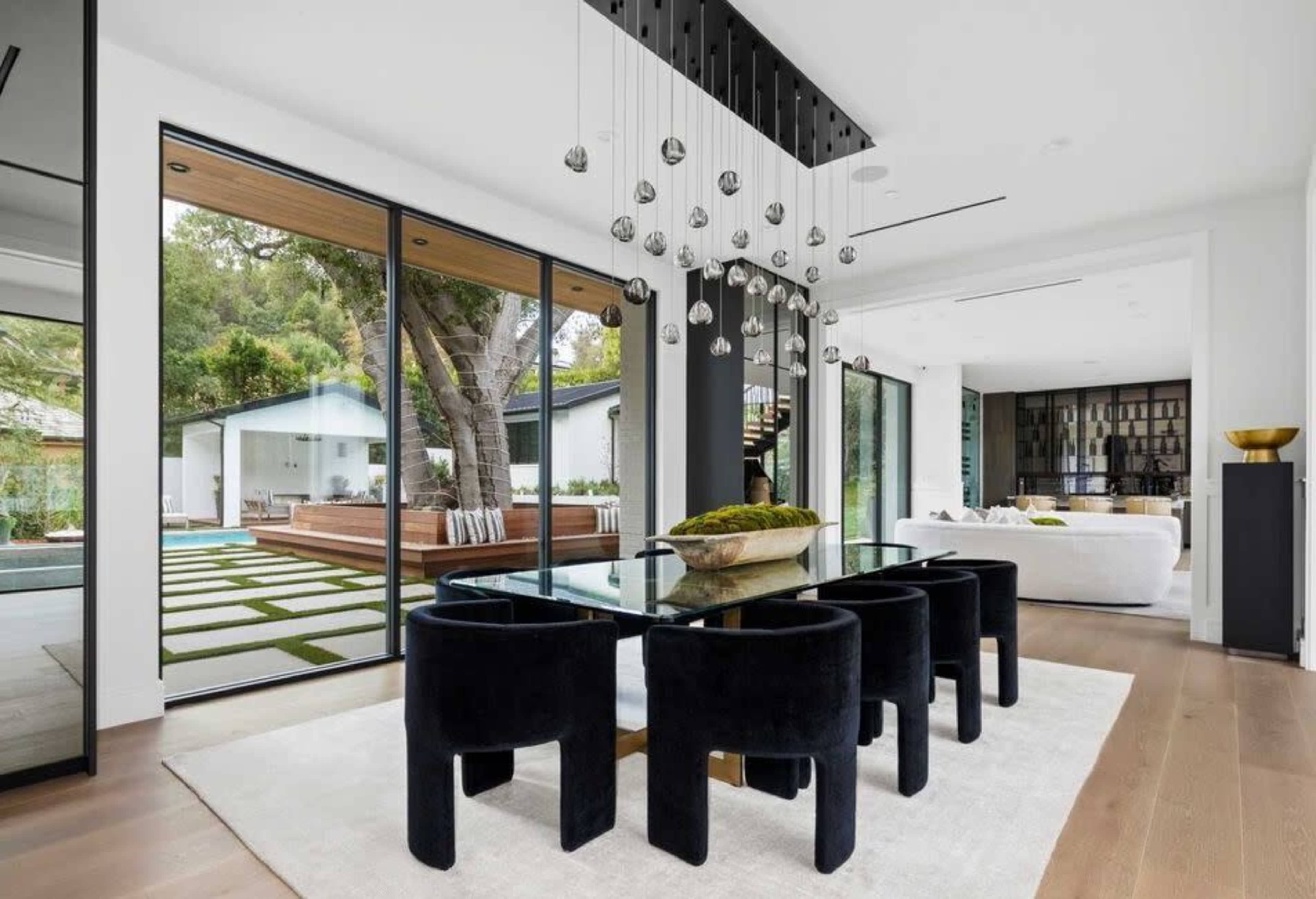 A modern dining area features a glass table surrounded by black chairs, with large windows providing a view of a landscaped outdoor space.