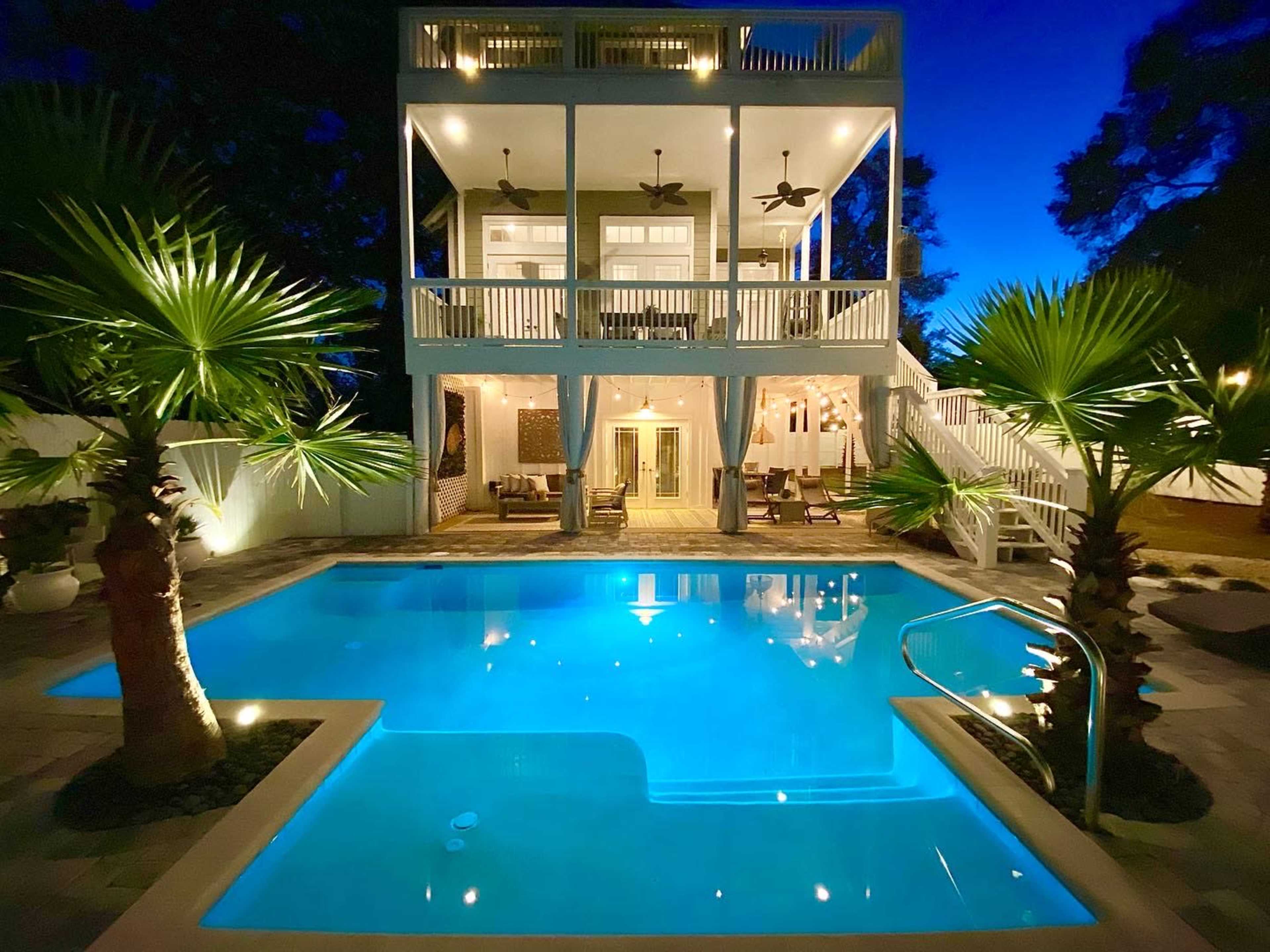 A two-story house with large balconies overlooks a illuminated swimming pool surrounded by palm trees at twilight.