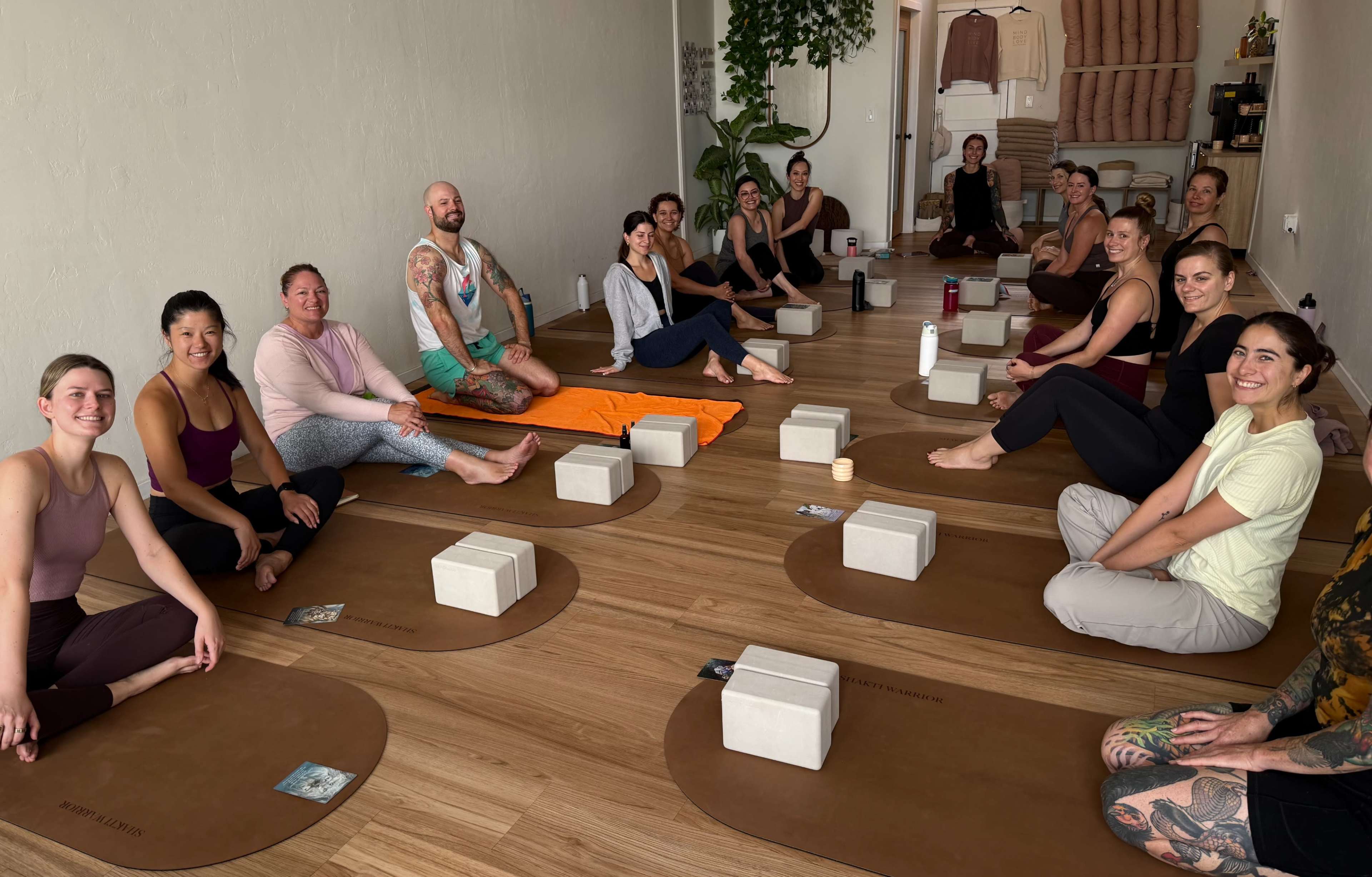 A group of individuals participate in a yoga class seated on mats in a well-lit studio with block props arranged in front of them.