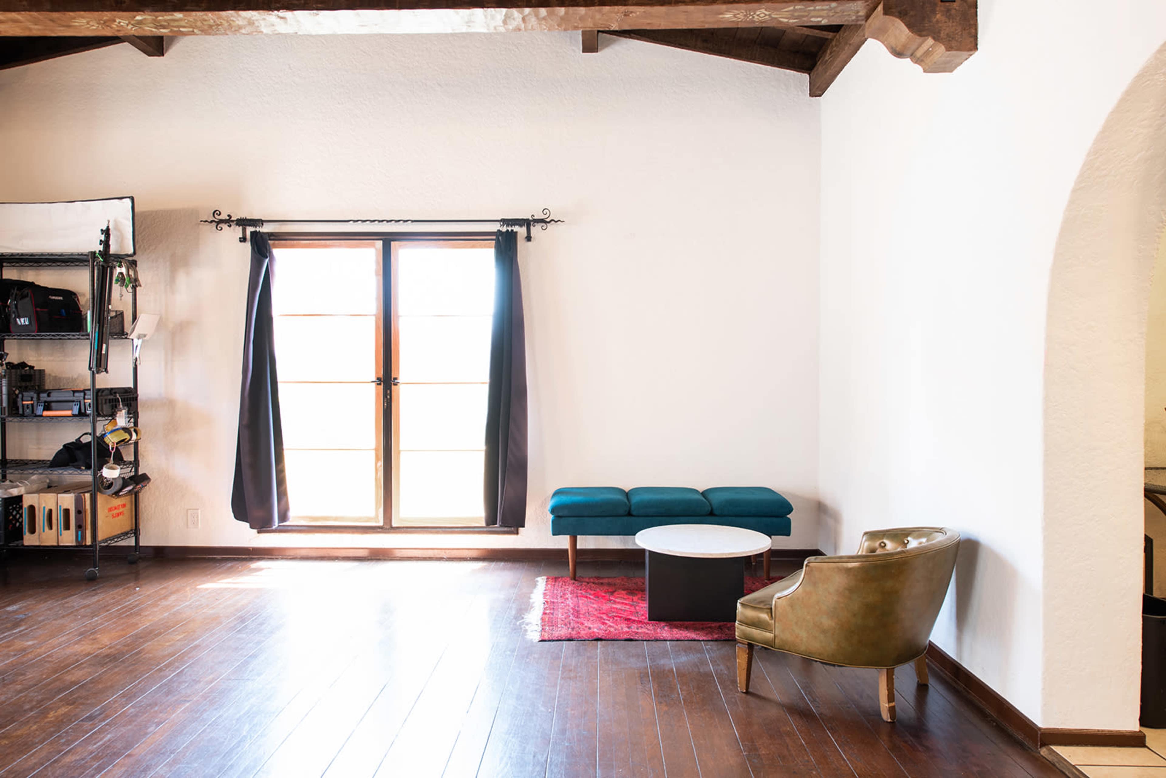 A spacious room with wooden floors, a large window with black curtains, a green chair, a blue bench, and a round coffee table on a red rug.