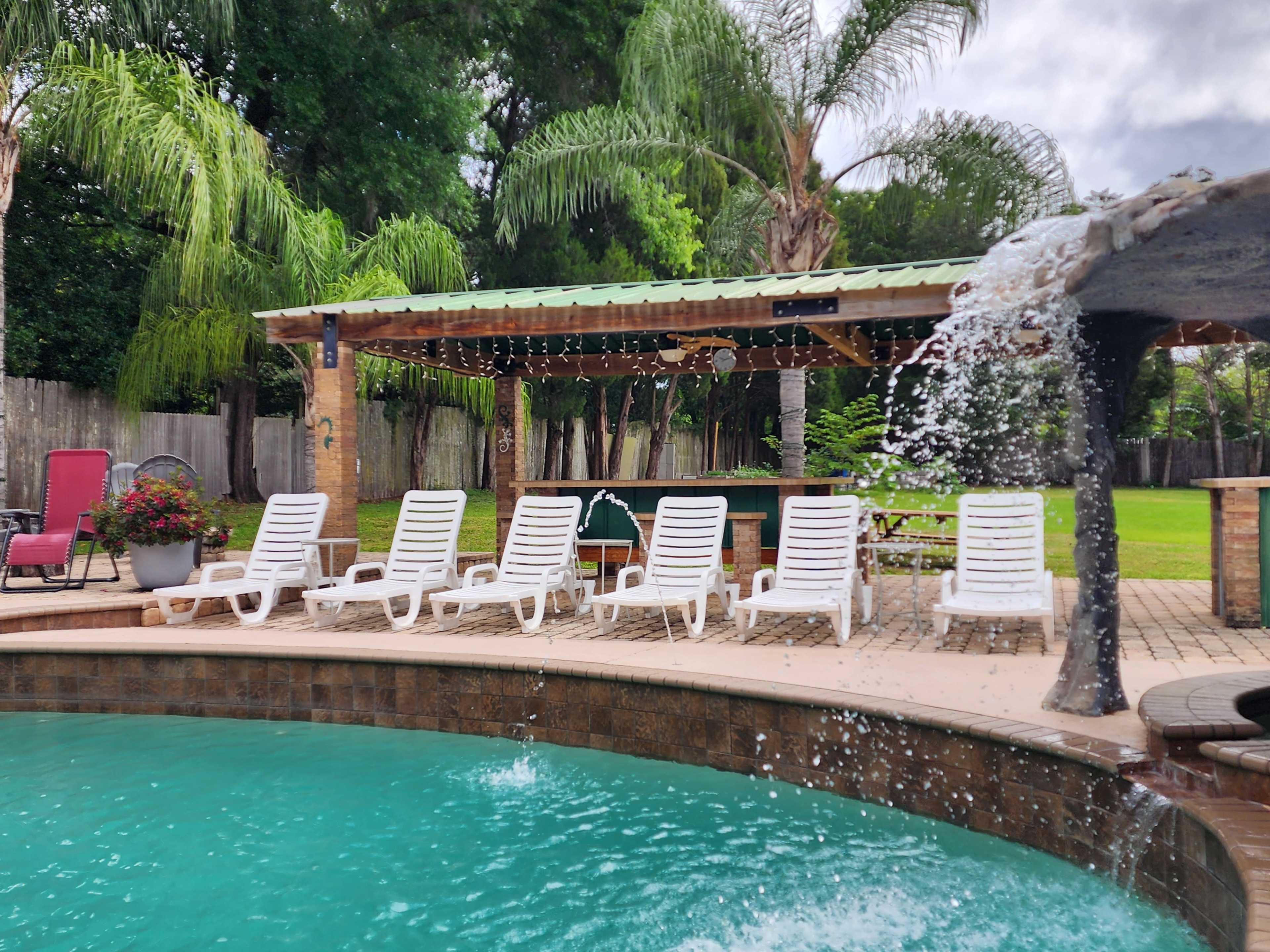A patio area with several white lounge chairs surrounds a swimming pool, featuring a stone waterfall and a pergola with string lights in a lush backyard.