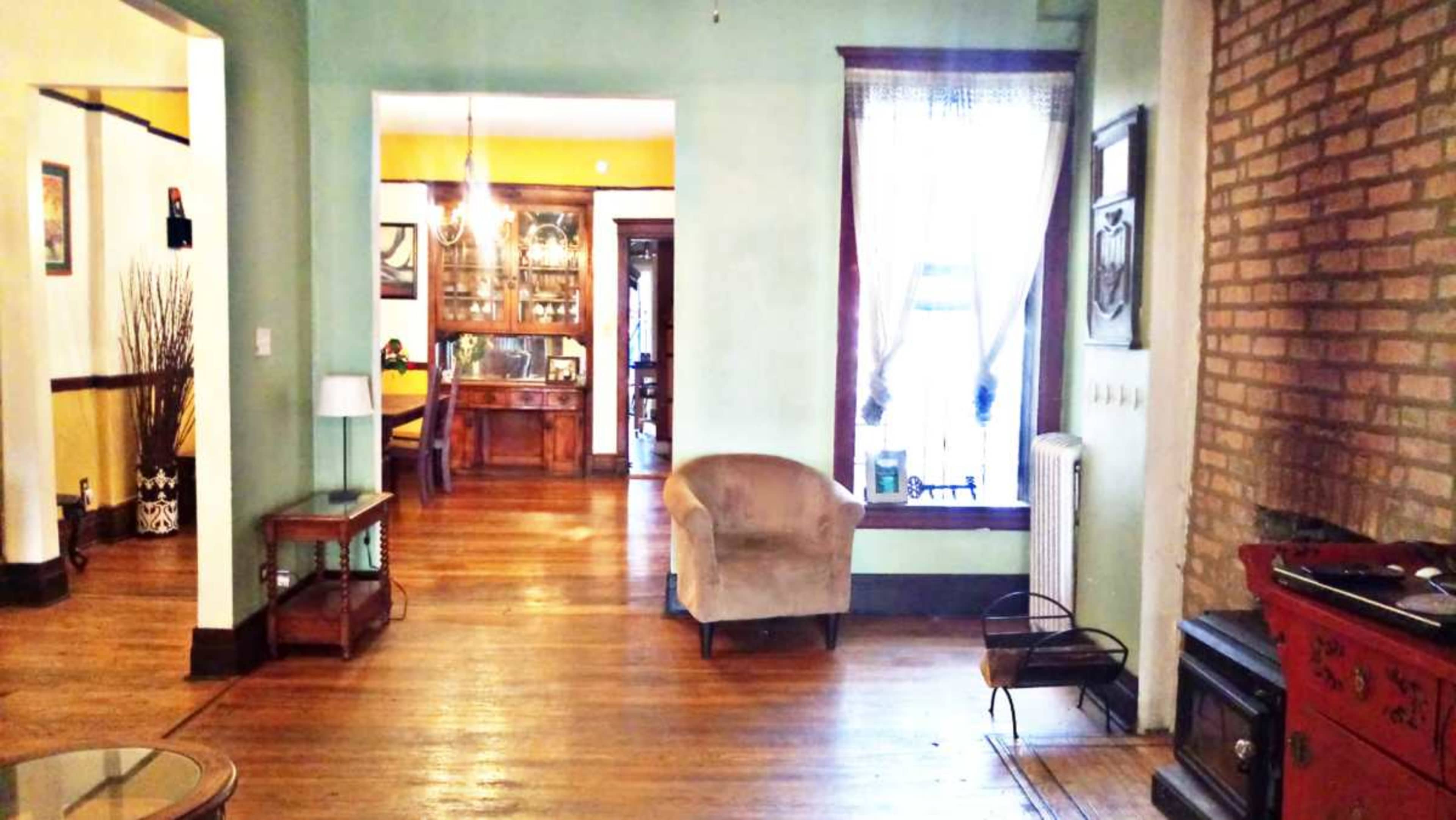 The room features a blend of wooden flooring and walls with a large window, a plush armchair, and a brick accent wall.