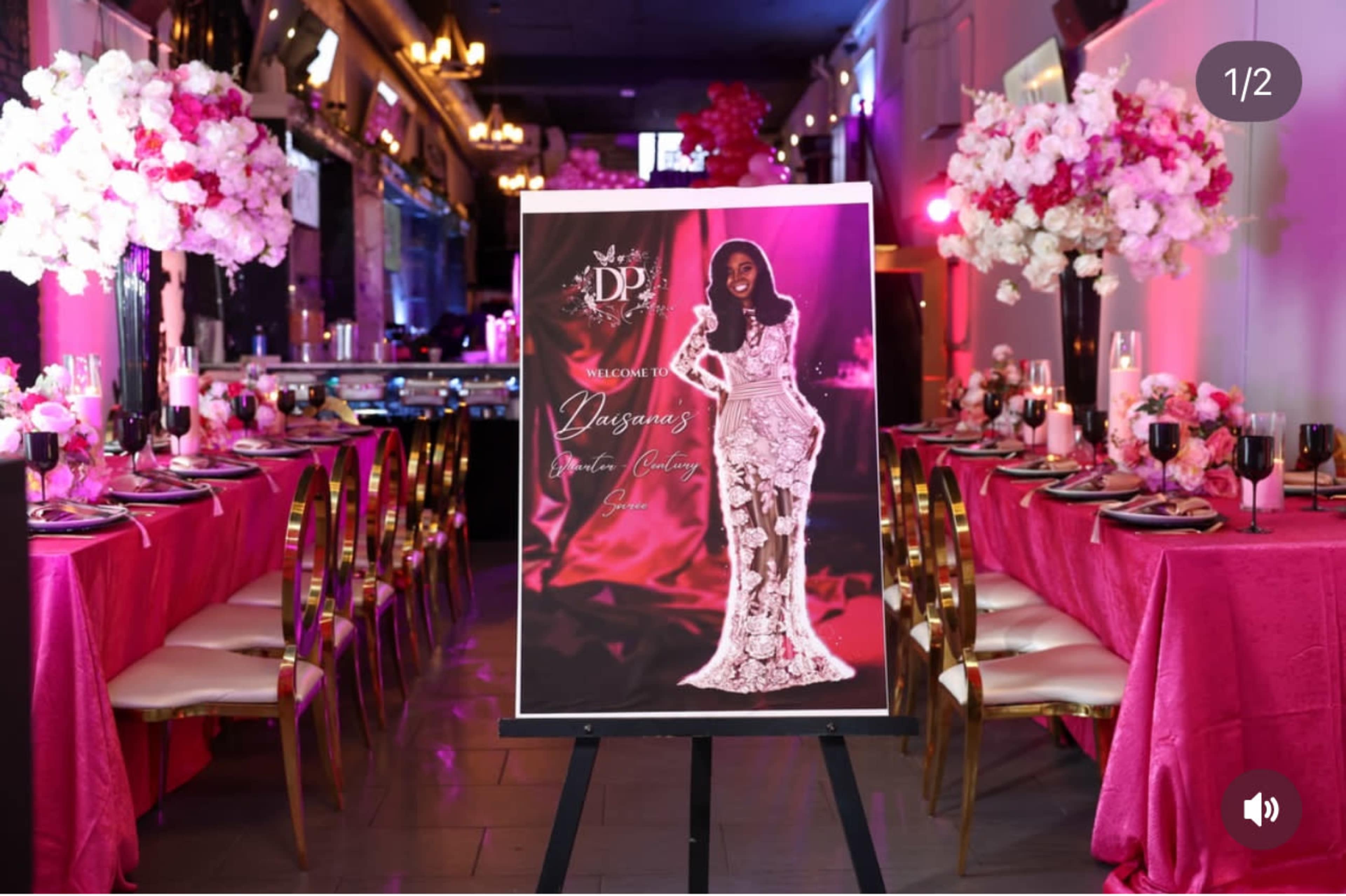 The image shows a decorated event space featuring elegantly set tables with pink tablecloths, floral centerpieces, and a large welcome sign showcasing a formal gown.