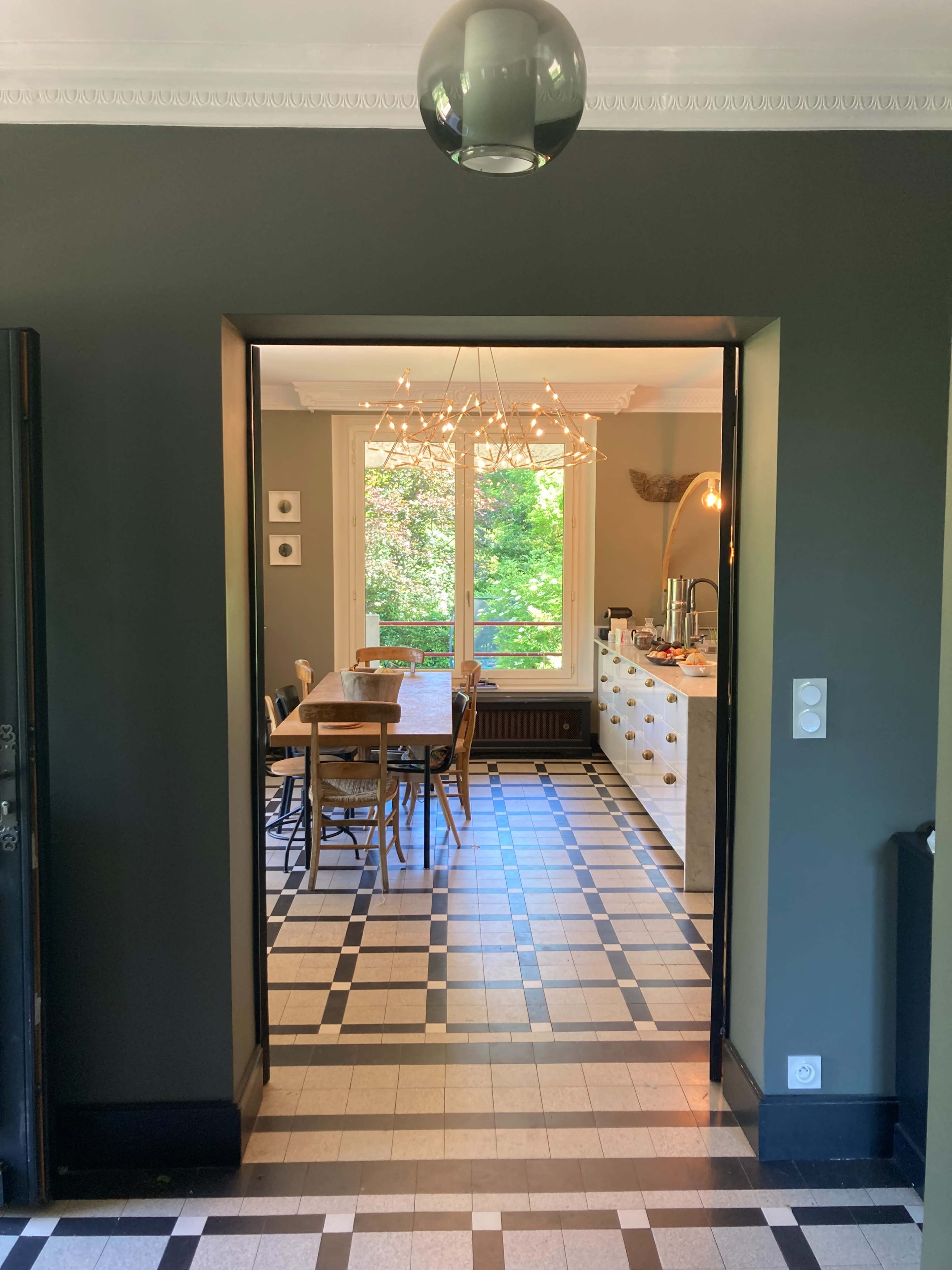 A doorway frames a dining area featuring a wooden table and chairs, illuminated by a chandelier, with a view of trees through a large window.