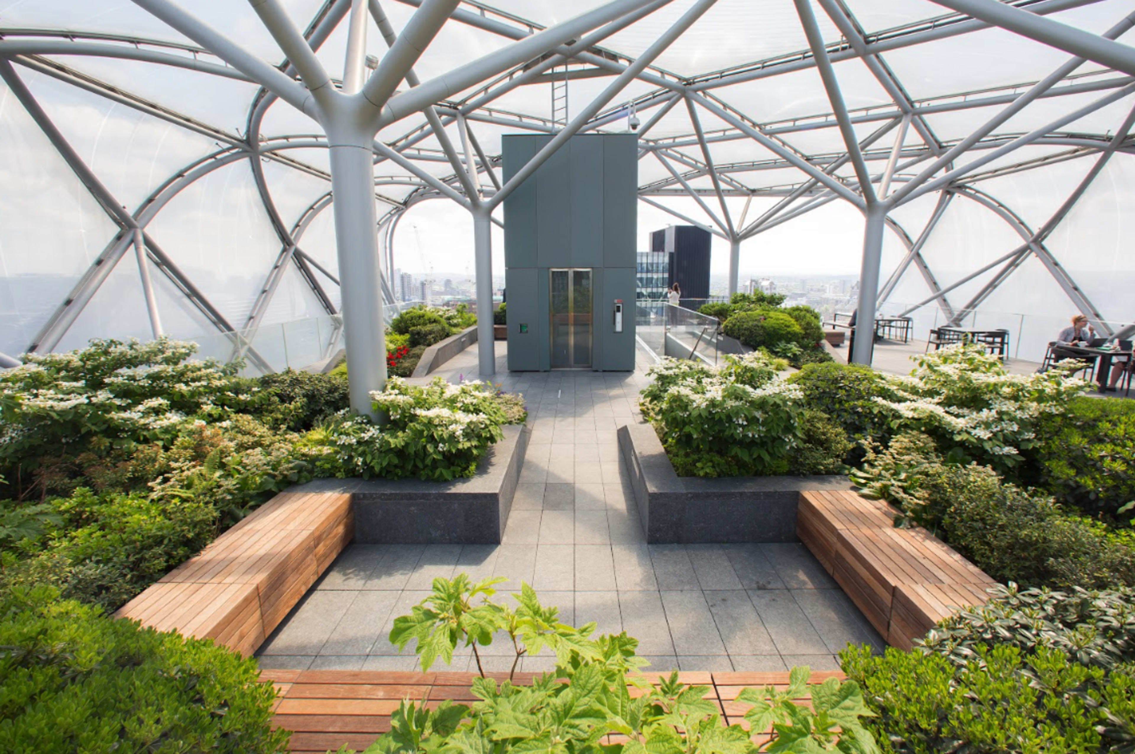 The image shows a modern rooftop garden featuring greenery, wooden benches, and a structure with a transparent canopy.