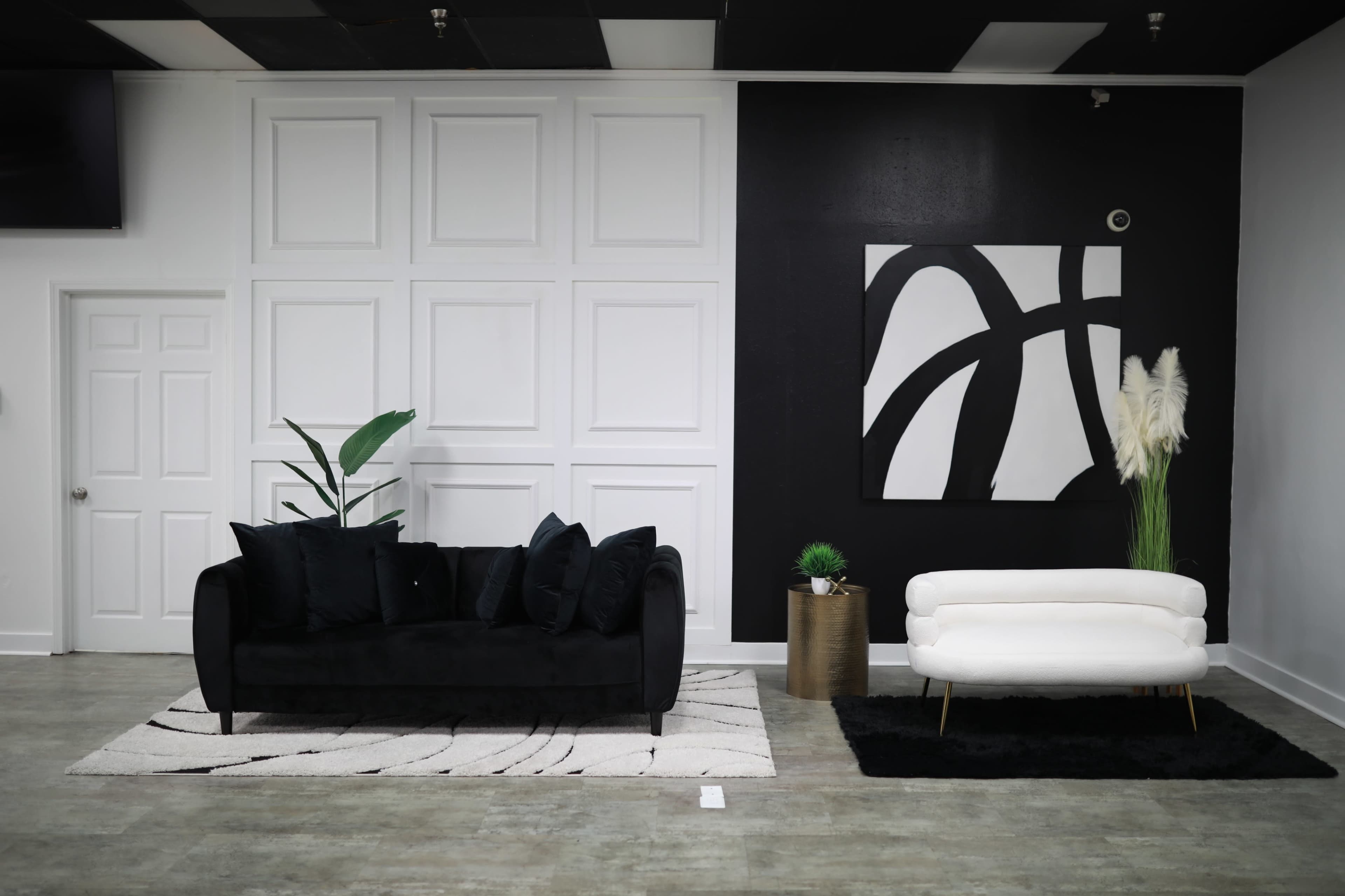 The image features a modern interior with a black sofa and a white loveseat, arranged on a patterned rug against a backdrop of white and black walls.