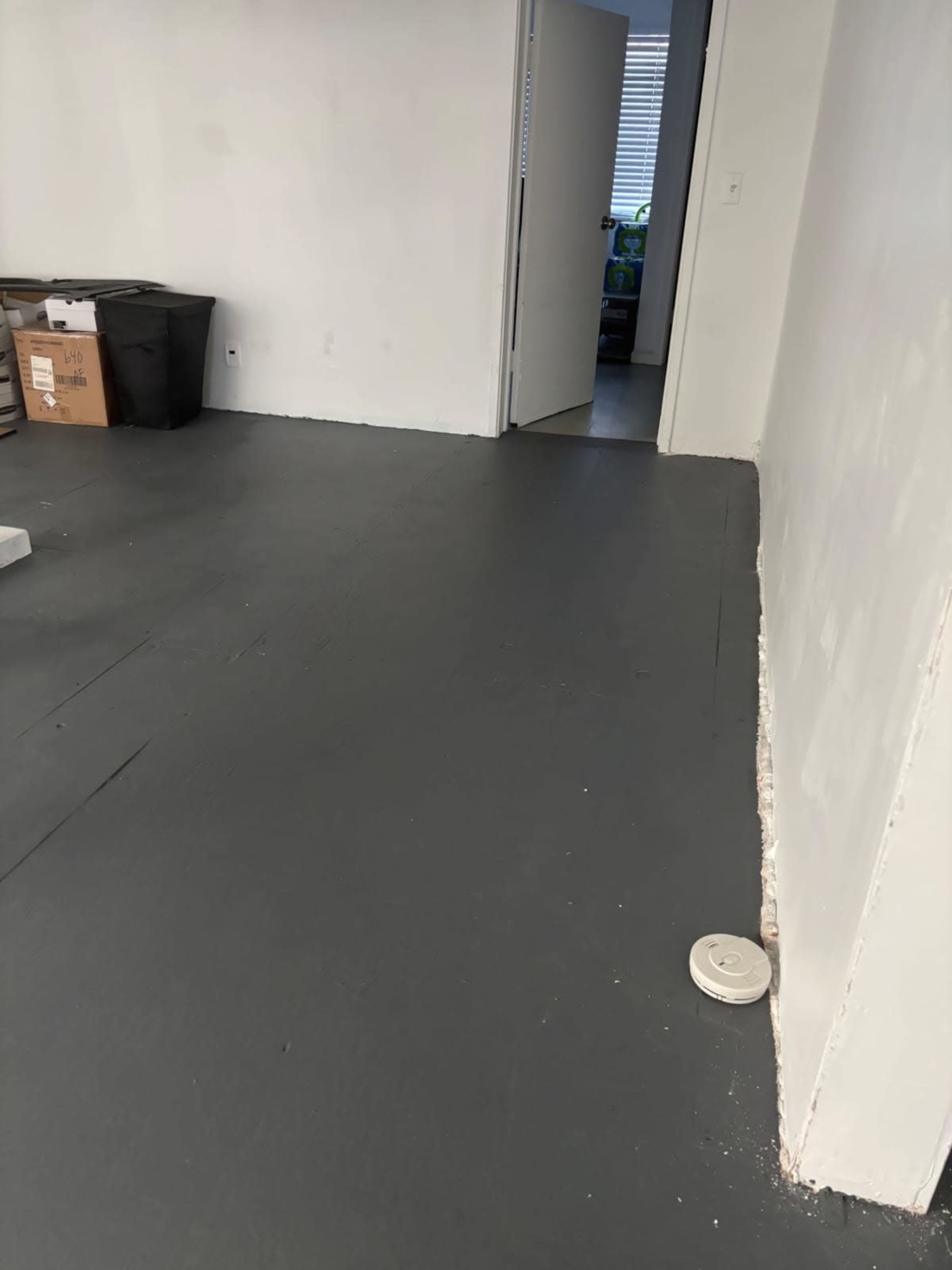 The image shows a sparsely furnished room with a gray floor, a doorway leading to another room, and boxes stacked in the corner.