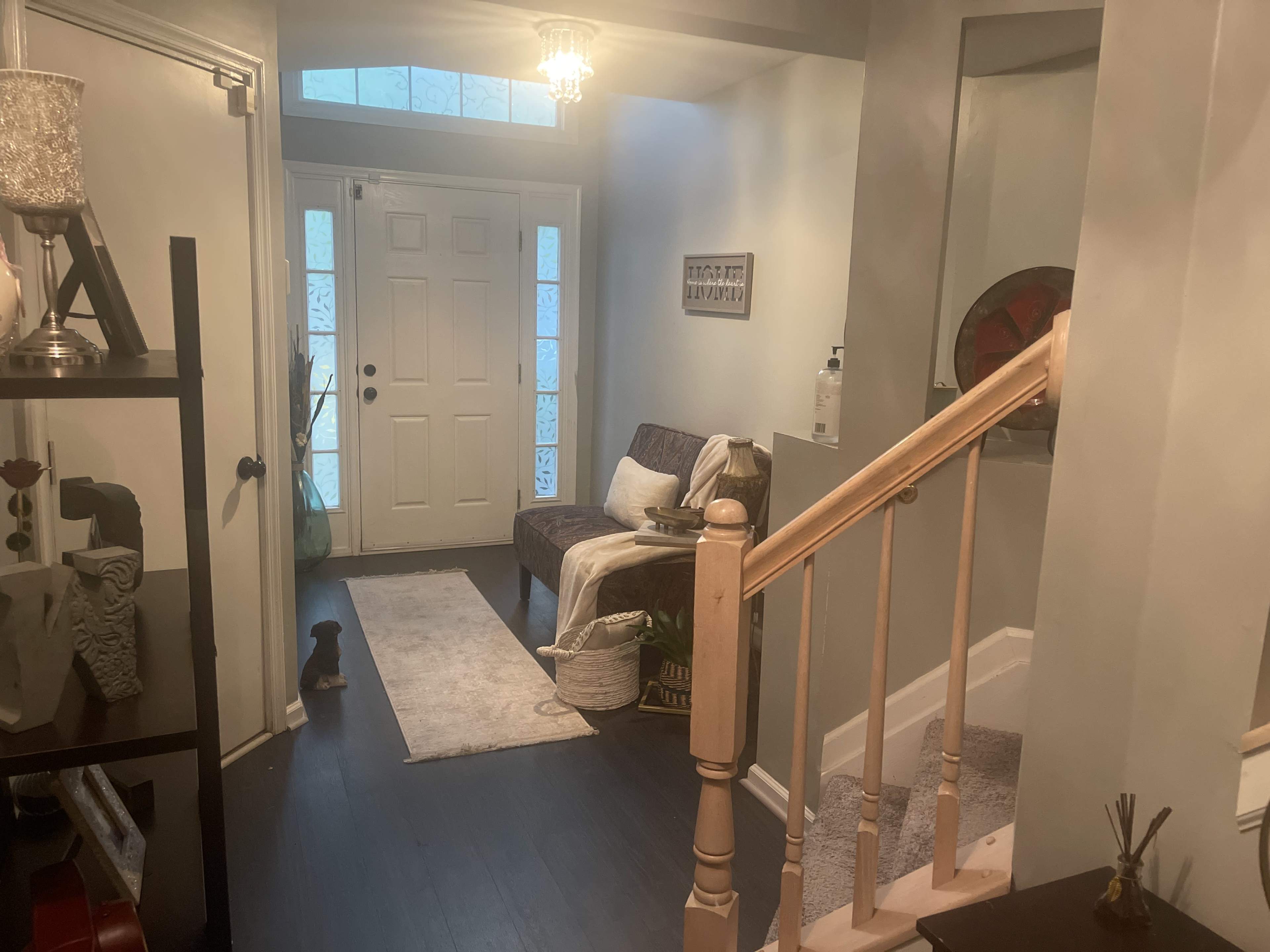 The image shows a hallway with a staircase on the right, a seating area with a throw blanket, and a view of the front door at the end.