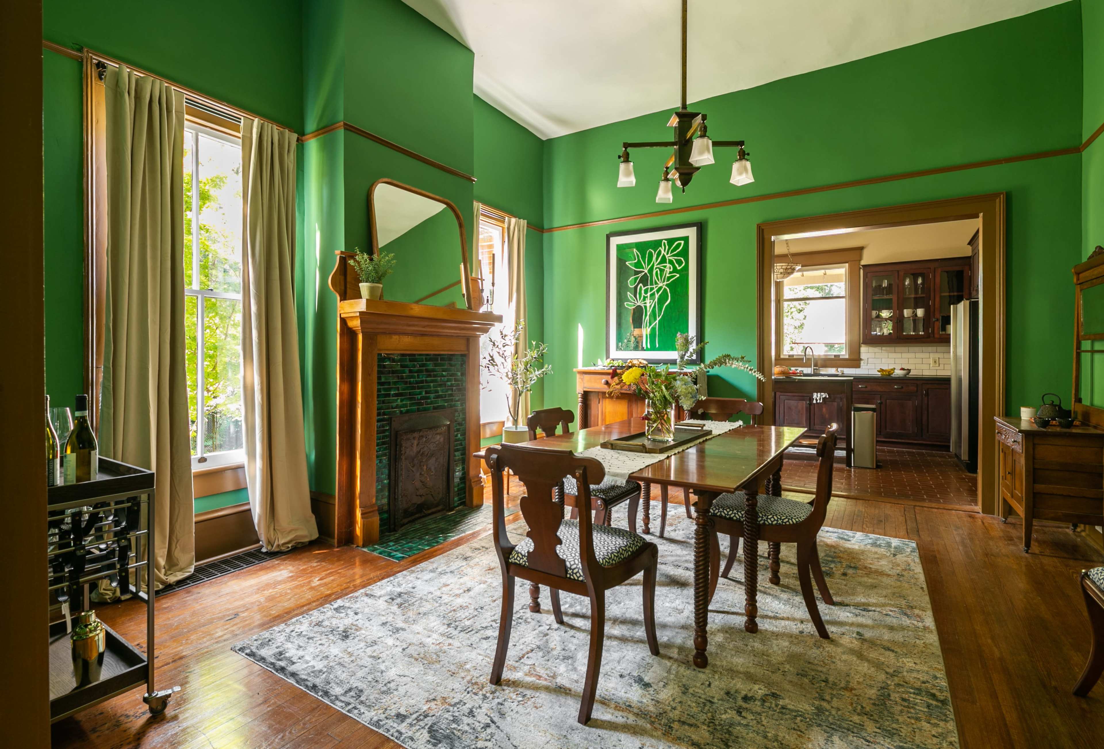 The room features green walls, a wooden dining table with four chairs, and a fireplace, with a view into a kitchen visible through an adjoining doorway.