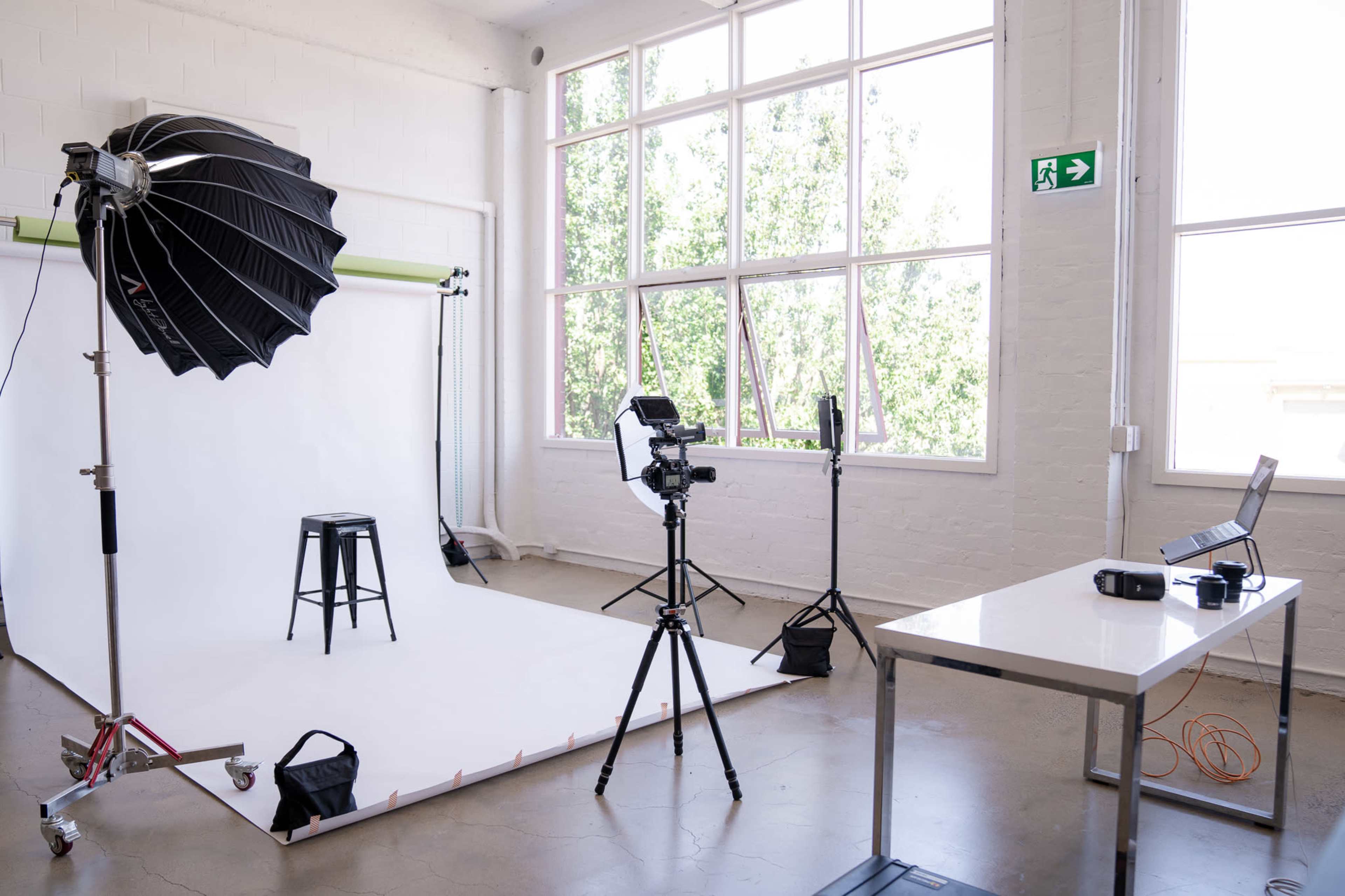 The image shows a photography studio setup with a white backdrop, a stool, various lighting equipment, and a camera on a tripod.