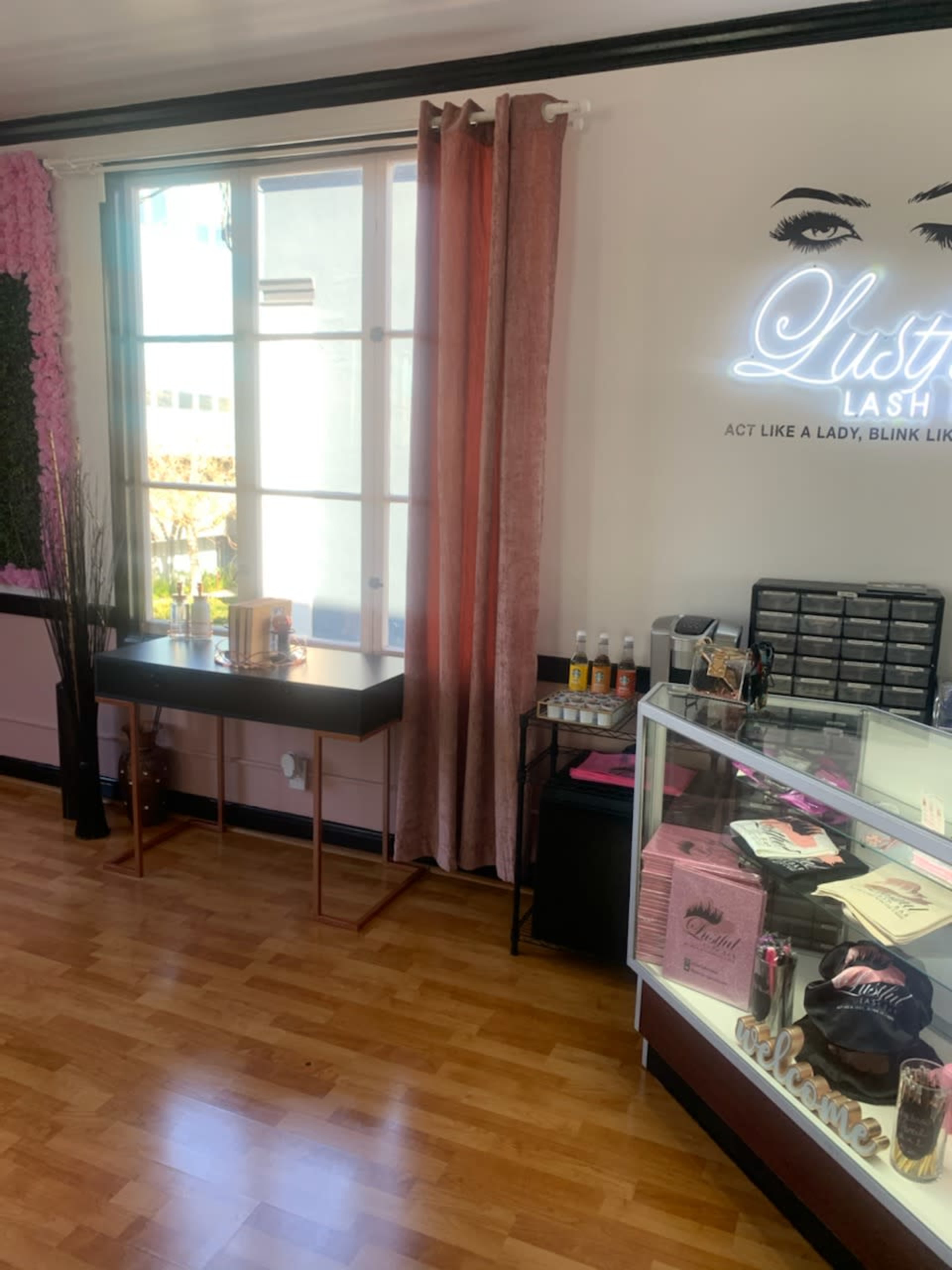 The image shows the interior of a lash studio featuring a display case, a table, and light pink curtains near a window.